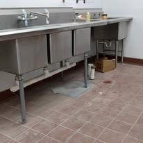Stainless steel sinks in a commercial setting, with plumbing visible and a brown tiled floor.