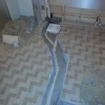 A herringbone-patterned floor with a trench cut through it, plumbing pipes visible, possibly under construction.