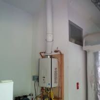 Water heater with vent pipe, mounted on a wall. Copper pipes and electrical components visible.