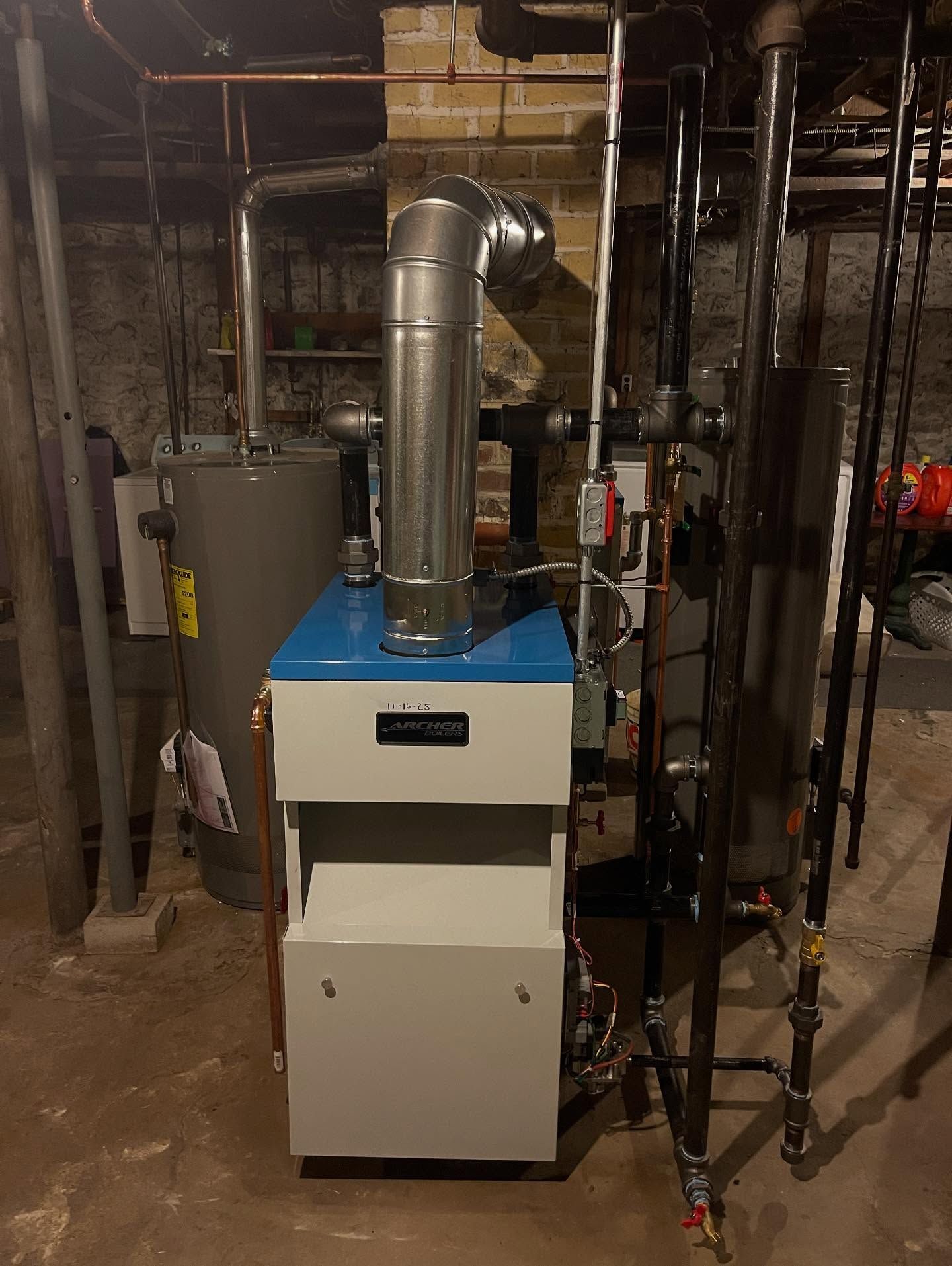 A beige and blue boiler sits in a basement with various black metal pipes, copper tubing, and a gray water heater.