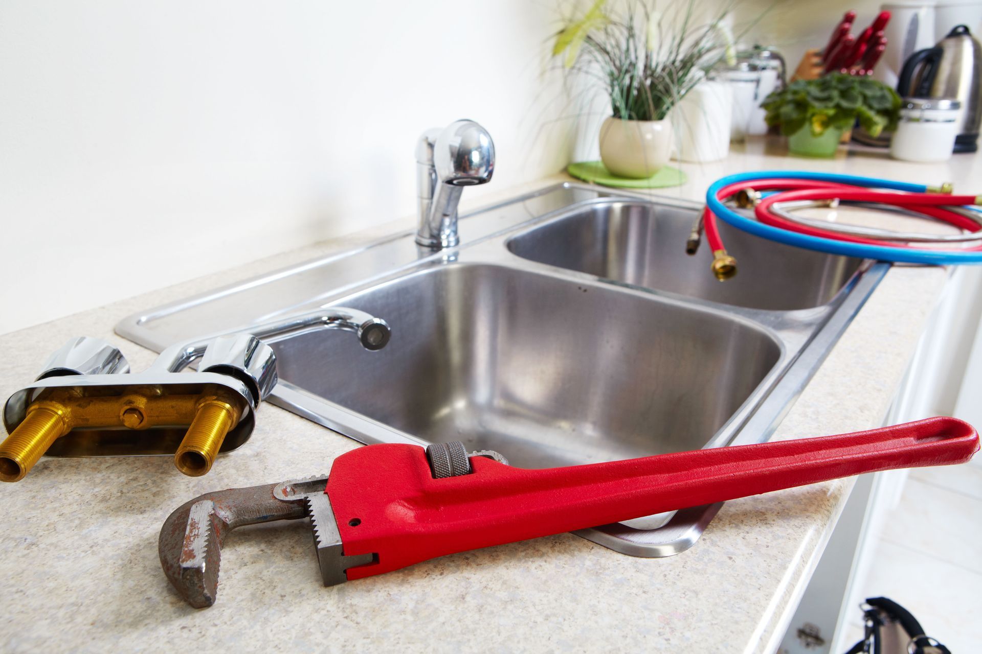 A red pipe wrench and a detached faucet fixture lie on a kitchen counter next to a double sink and coiled hoses.