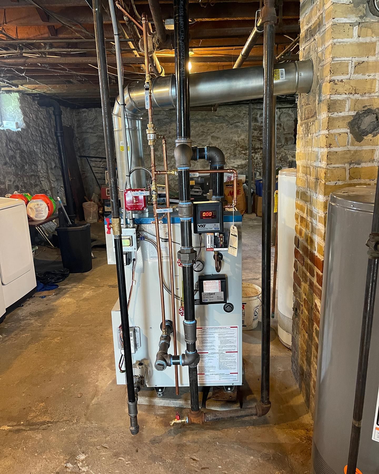 A residential boiler and metal piping system located in a basement with exposed brick and stone walls.