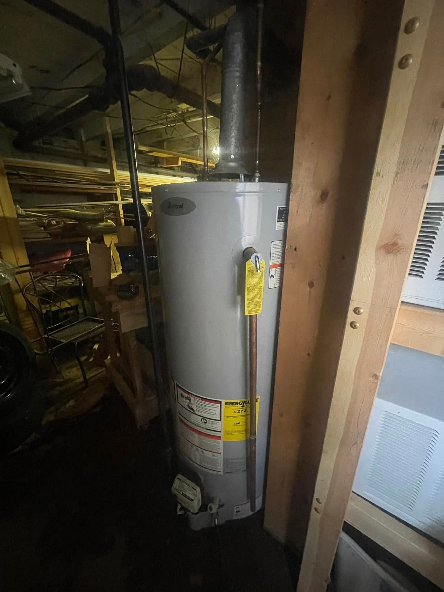 A tall, grey, cylindrical residential water heater installed in an unfinished basement next to wooden wall studs.