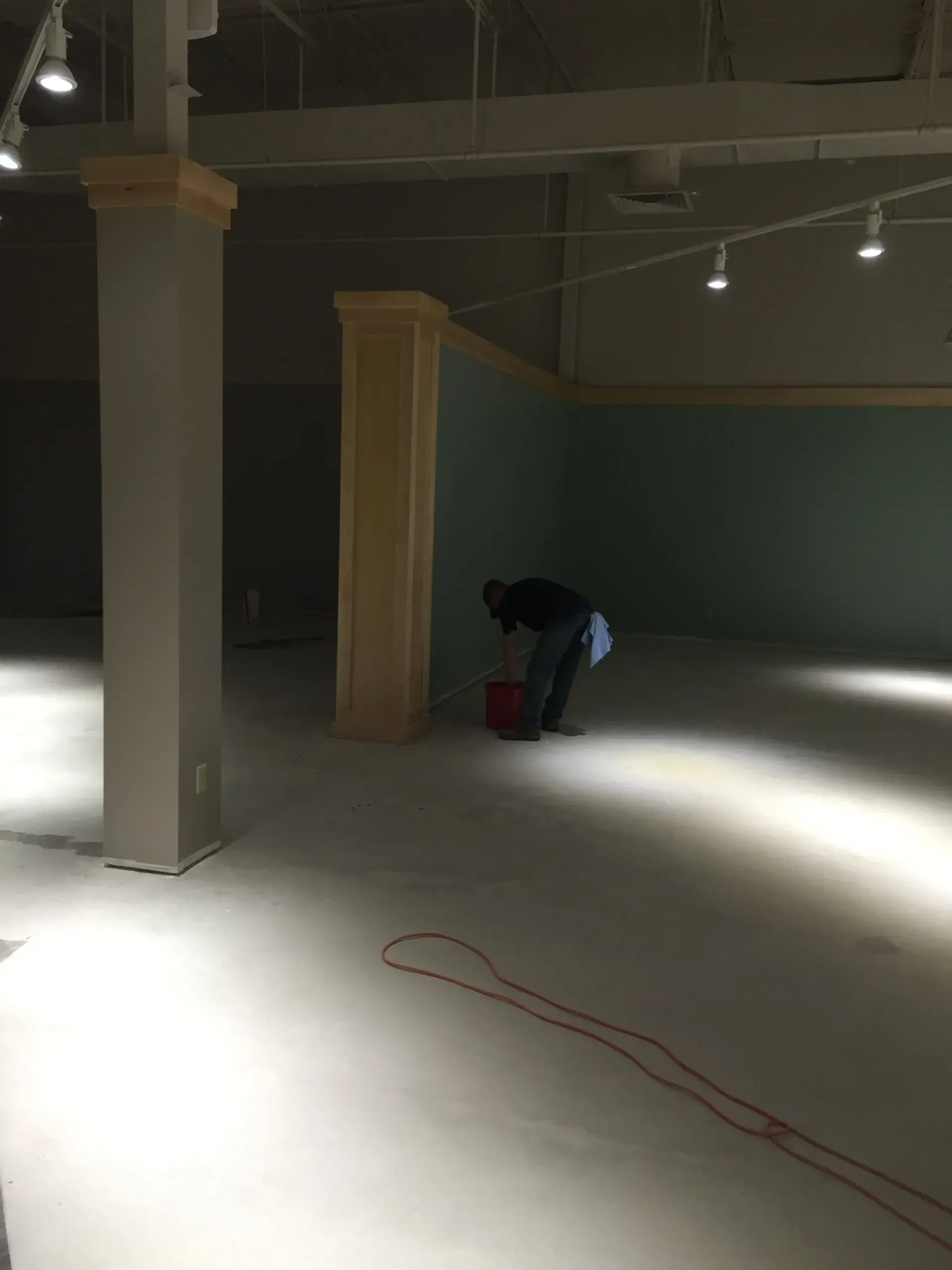 Person in empty room, painting floor. Beige walls, pillars. Red electrical cord.