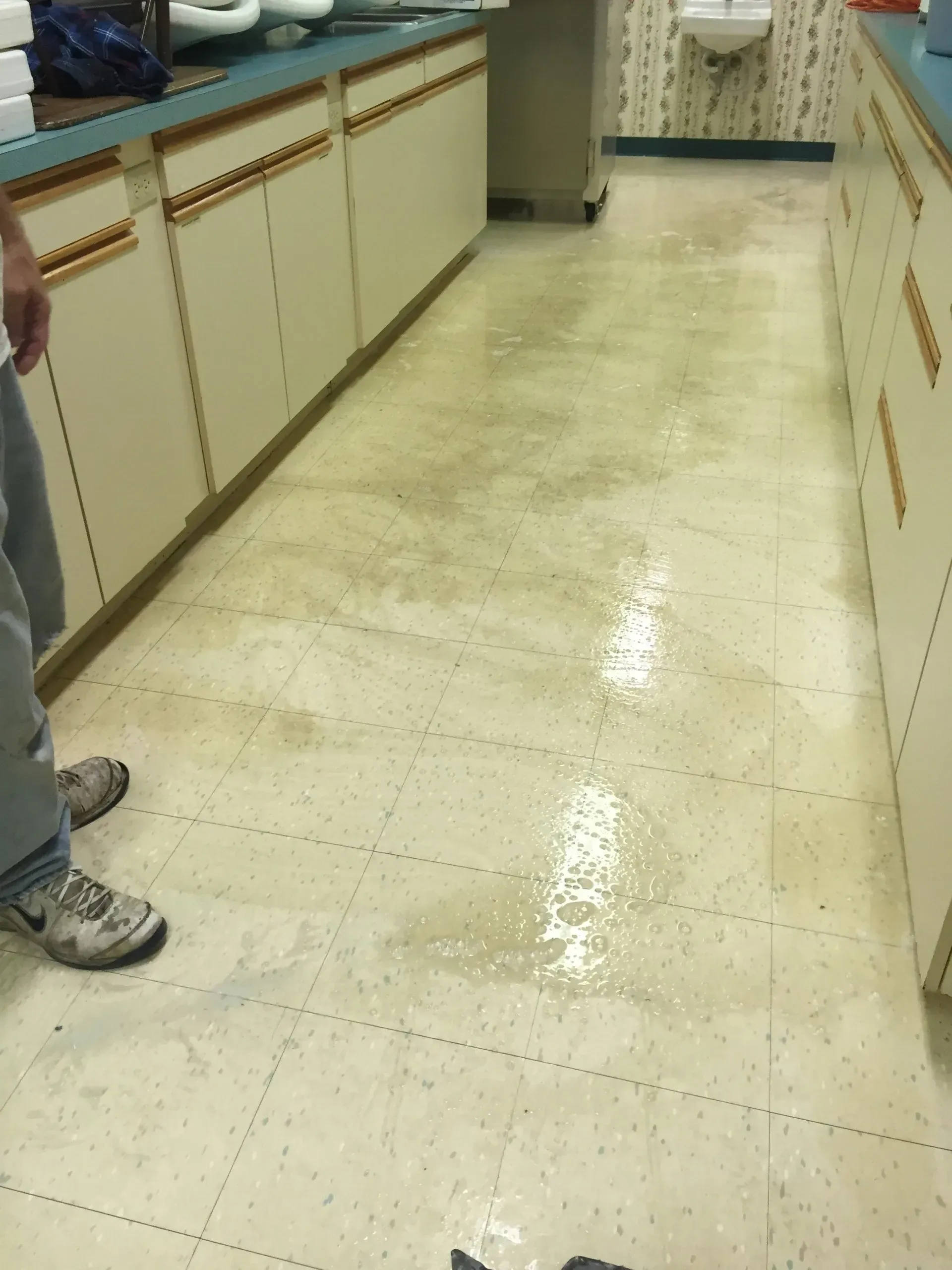 Wet, discolored floor in a kitchen with white cabinets. A person in jeans and sneakers stands nearby.