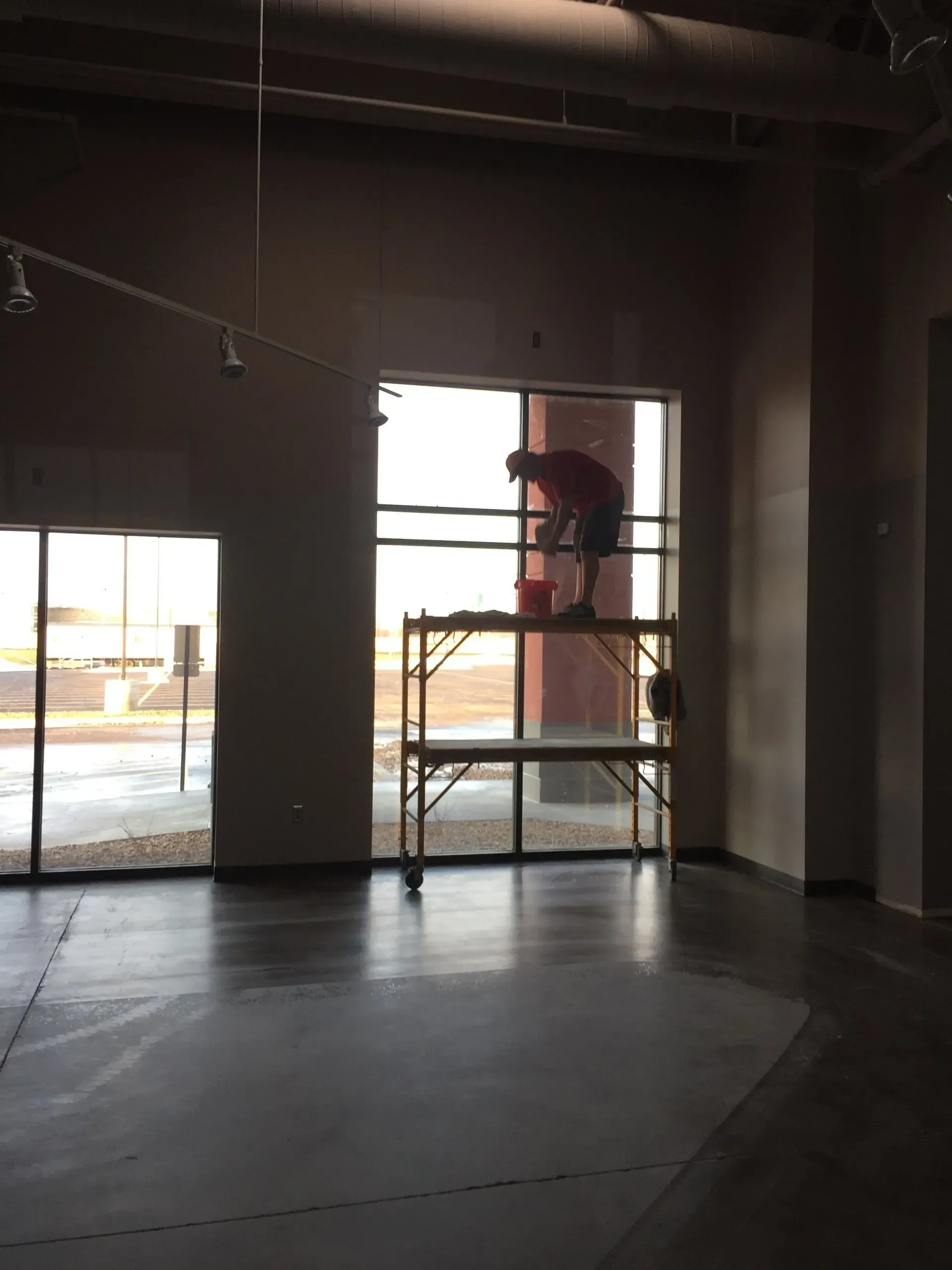 Person on scaffolding painting window in a room with concrete floor and large windows.