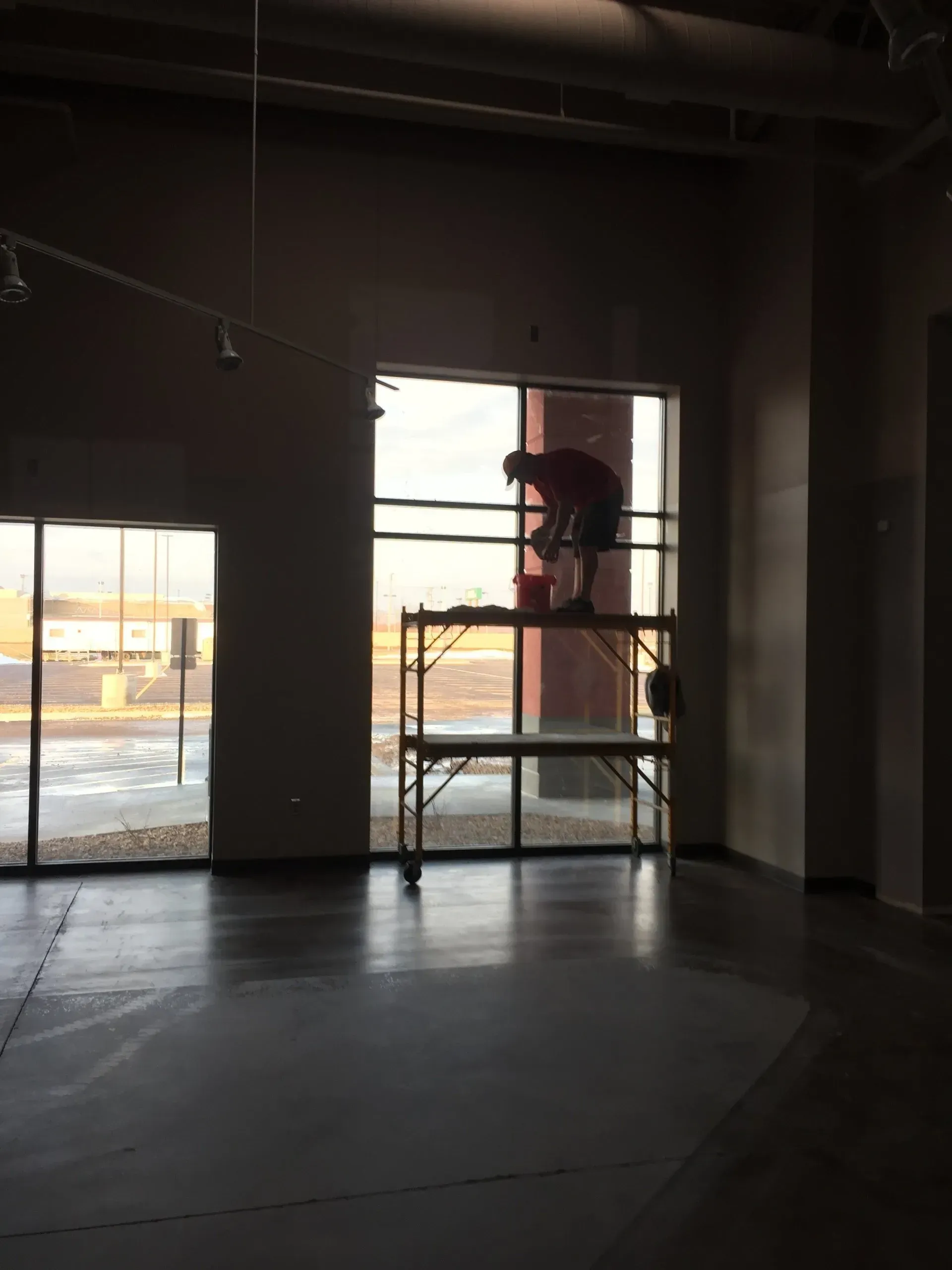 Person washing a large window, standing on a metal scaffold, inside a building with large windows and a dark floor.