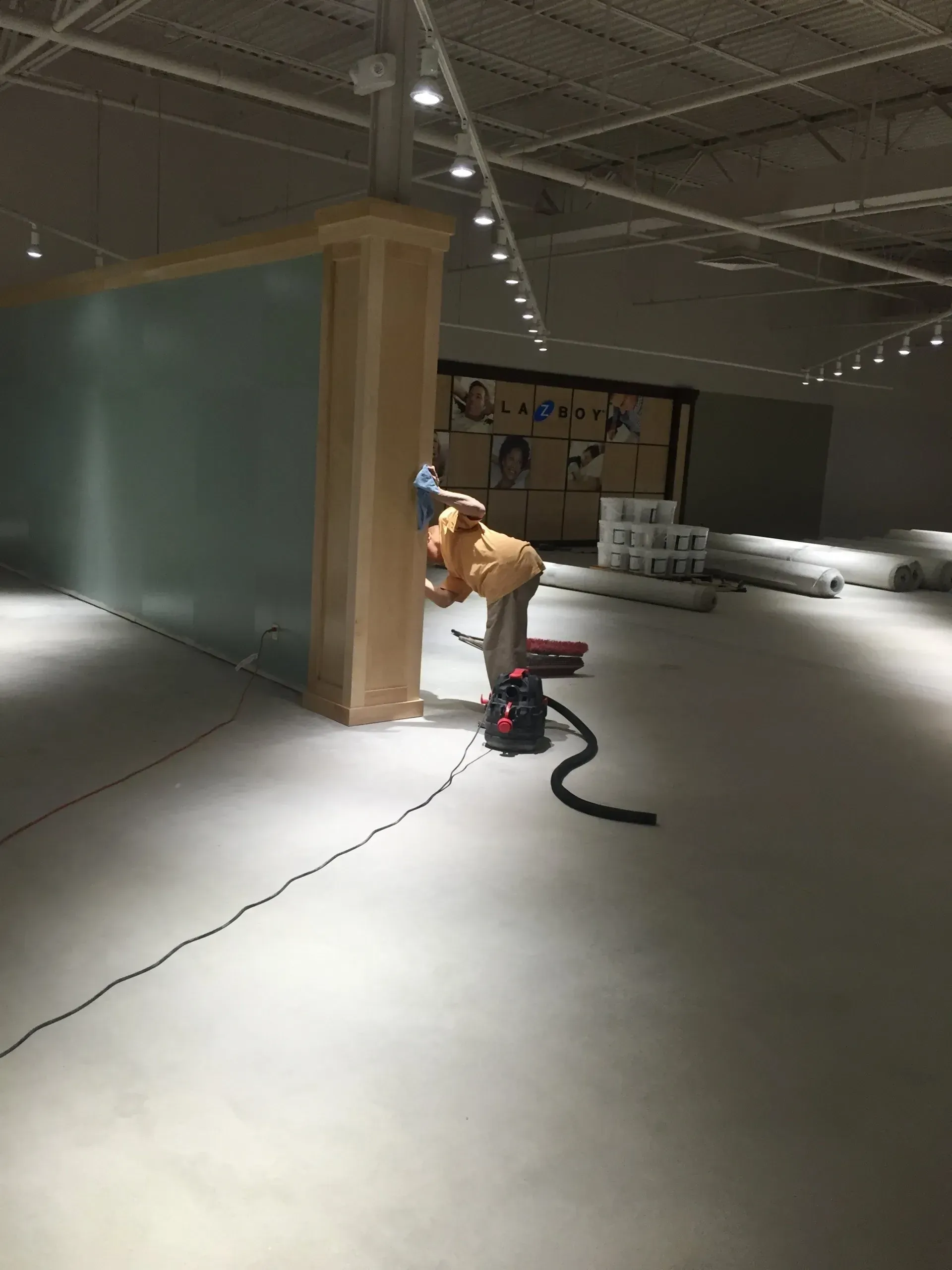 A person sanding a wood column in a large, empty room. Black vacuum hose on the floor.
