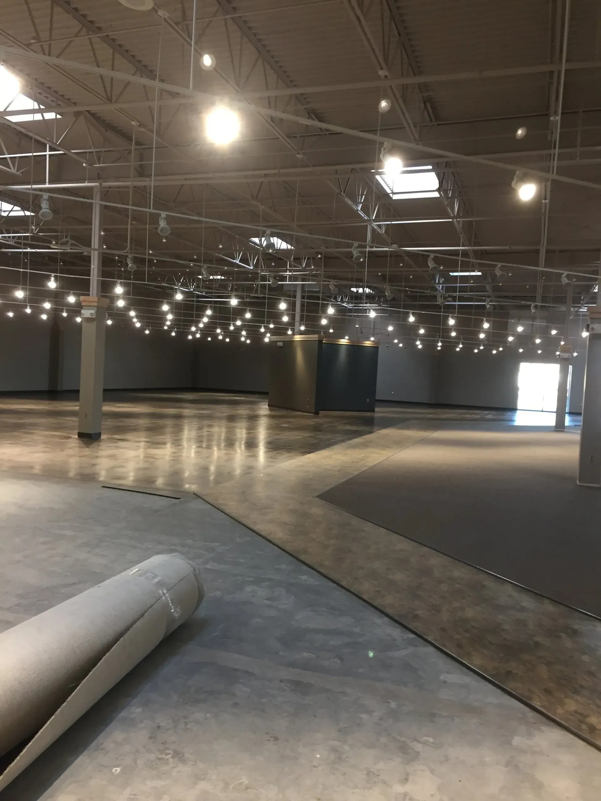 Large empty warehouse with concrete floor and many overhead lights. A roll of carpet sits on the floor.