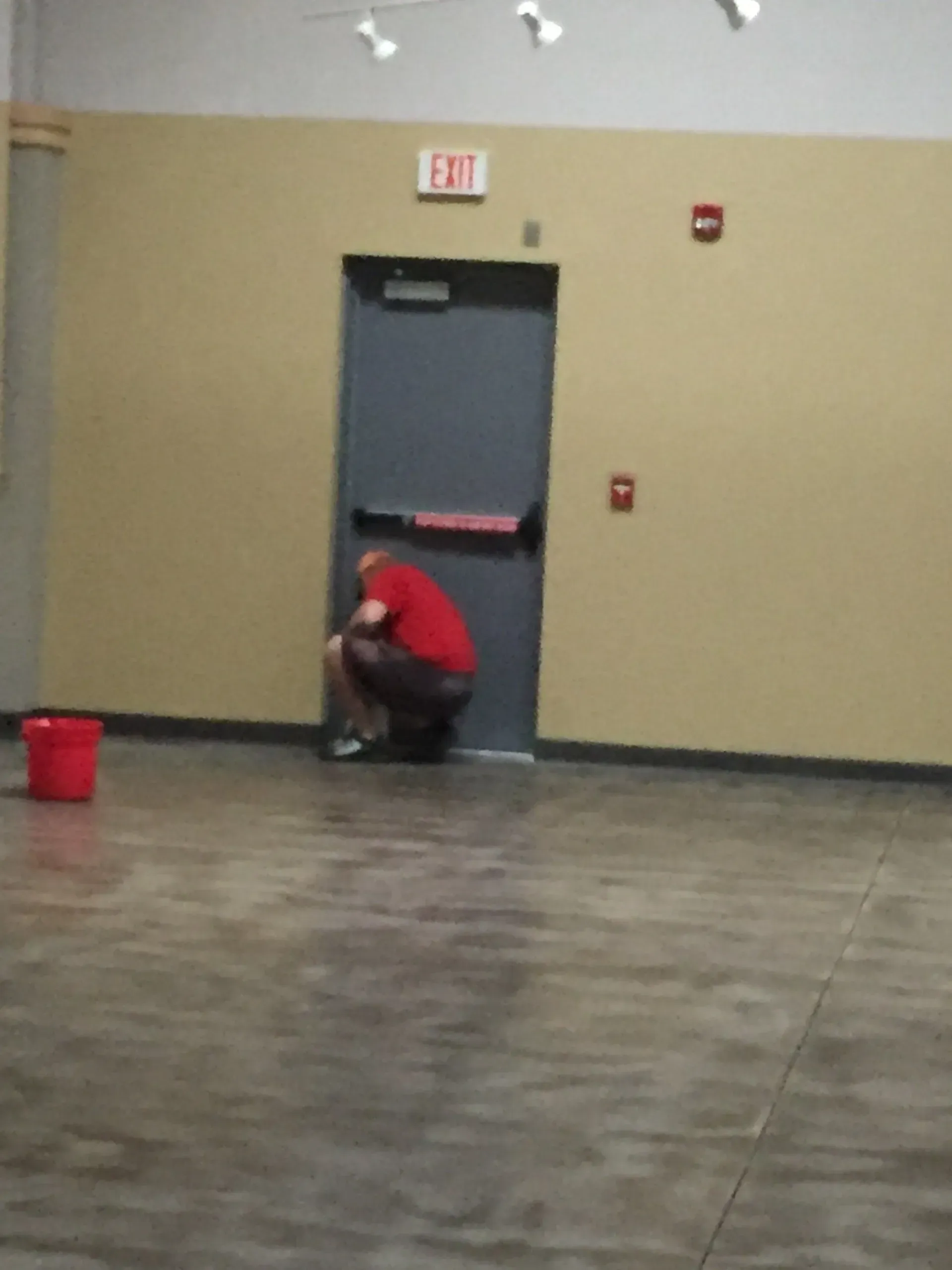 Person squatting near a closed gray exit door with a red exit sign. A red bucket sits on the floor.