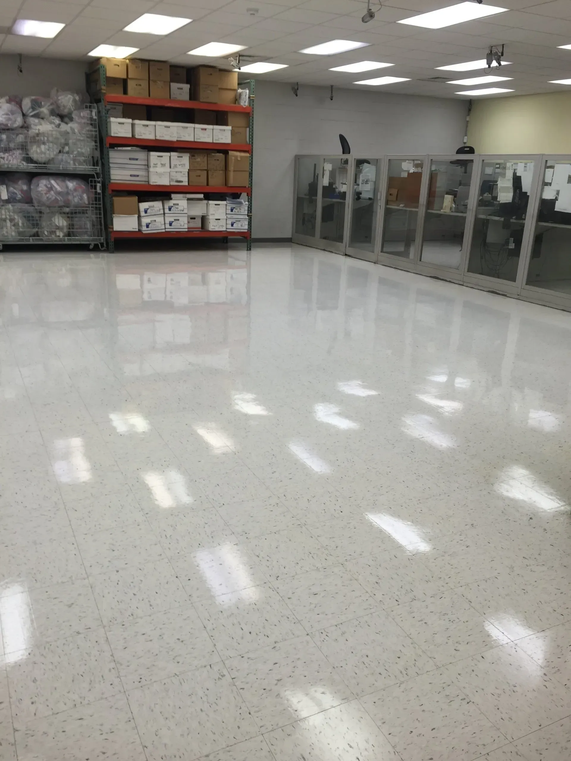 Bright, empty room with a glossy white floor. Shelves of boxes and cubicle walls are visible.