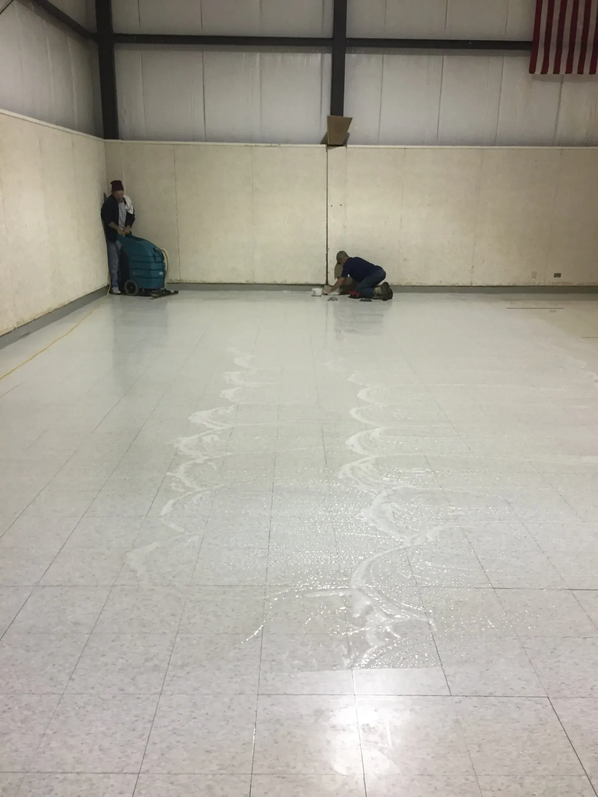Two people cleaning a large, tiled floor in a gymnasium. One operates a machine, the other scrubs.