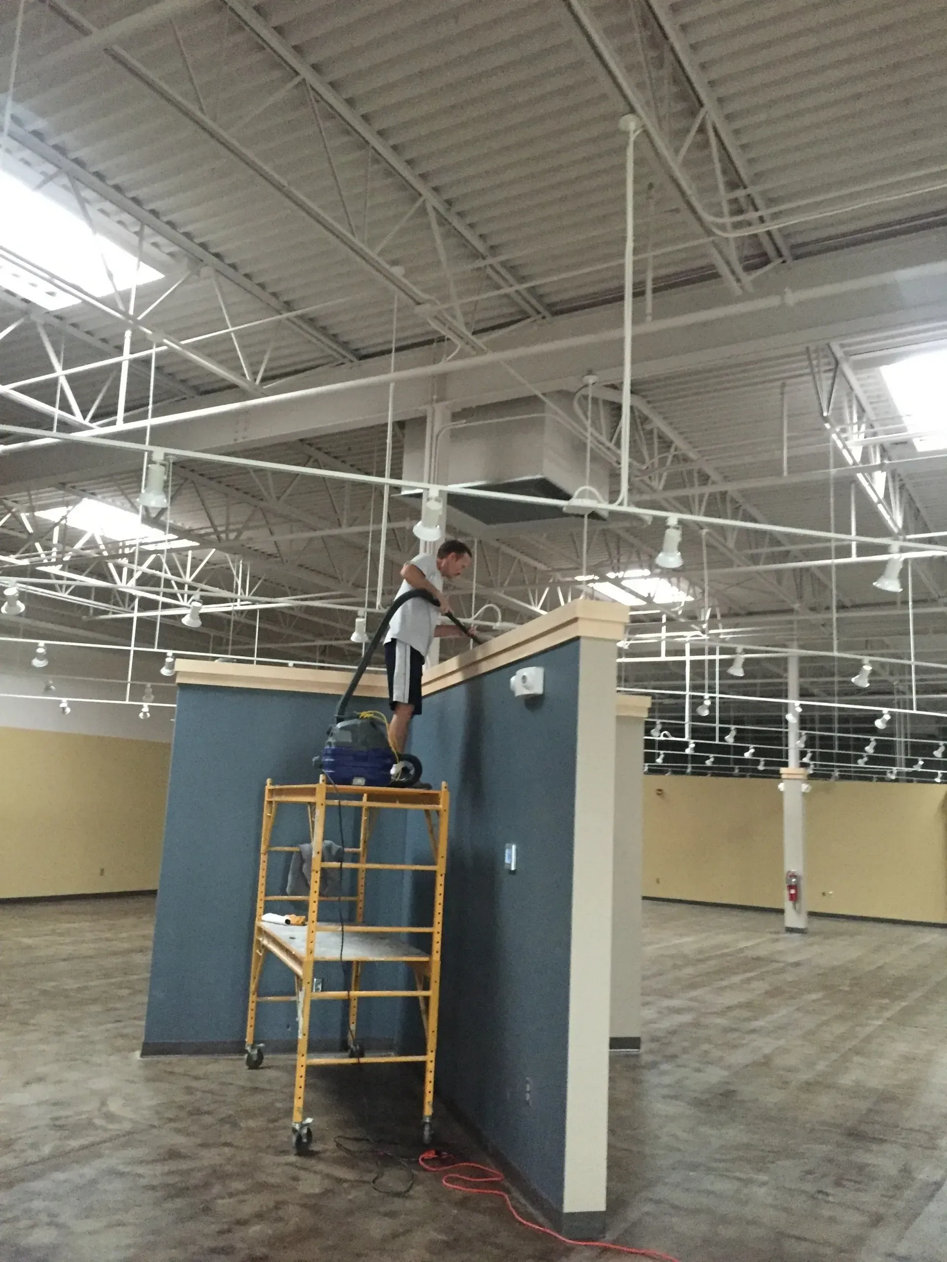 Man on a scaffold vacuums a wall in an unfinished, brightly lit retail space.