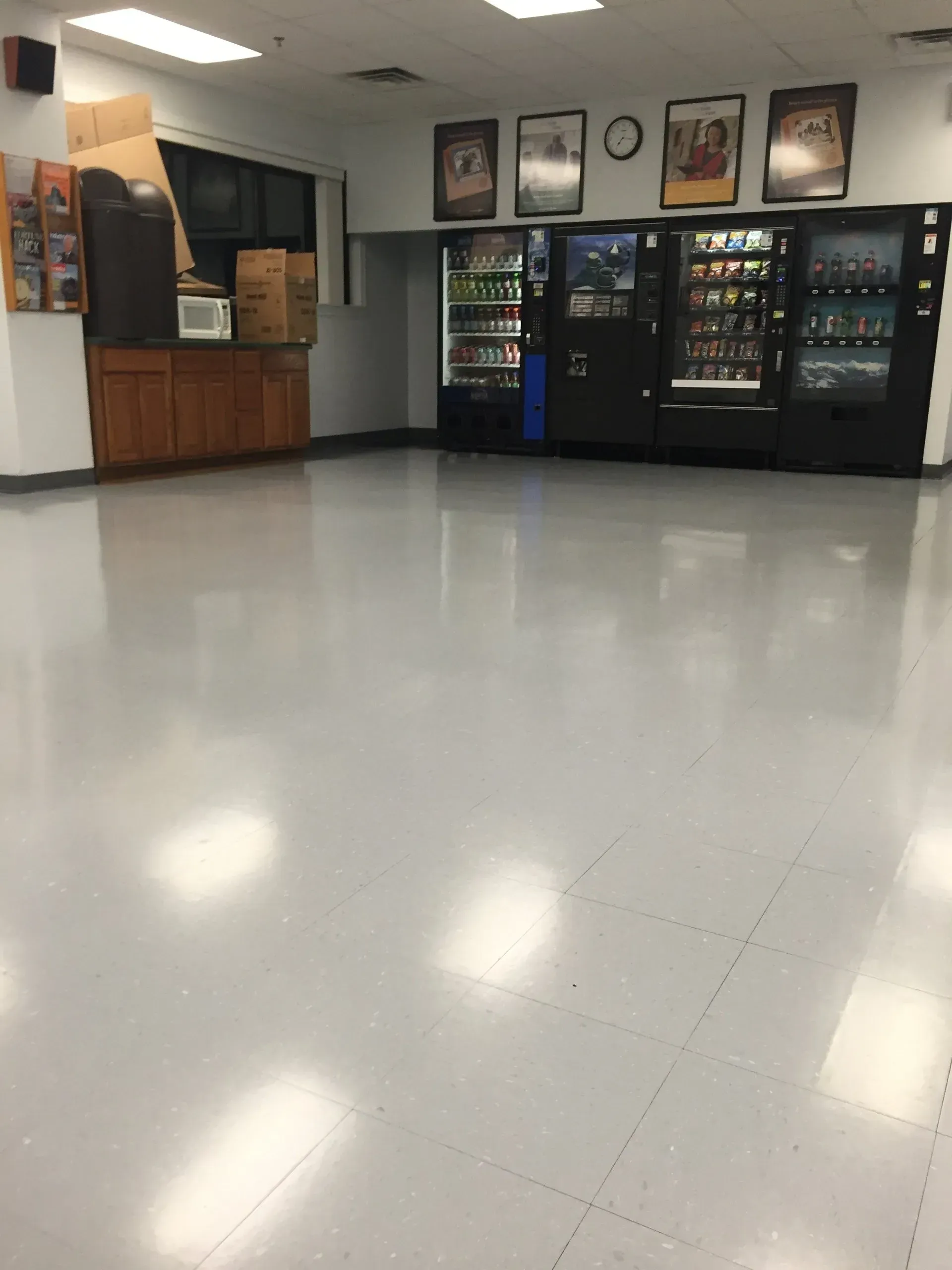 Empty room with vending machines, cabinets, and posters; gray floor and bright overhead lights.