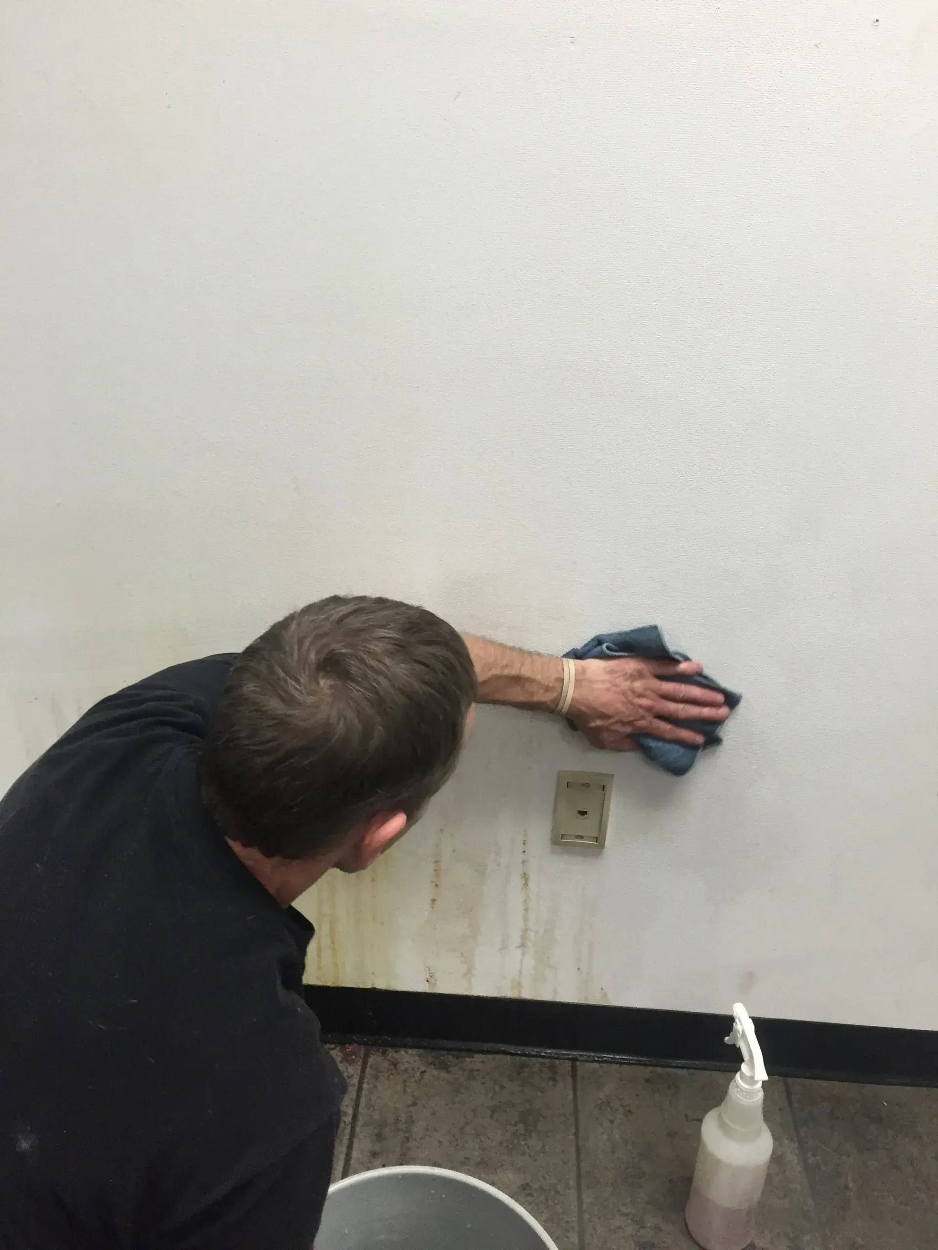 Person cleaning a white wall with a blue cloth near an electrical outlet and a black baseboard.