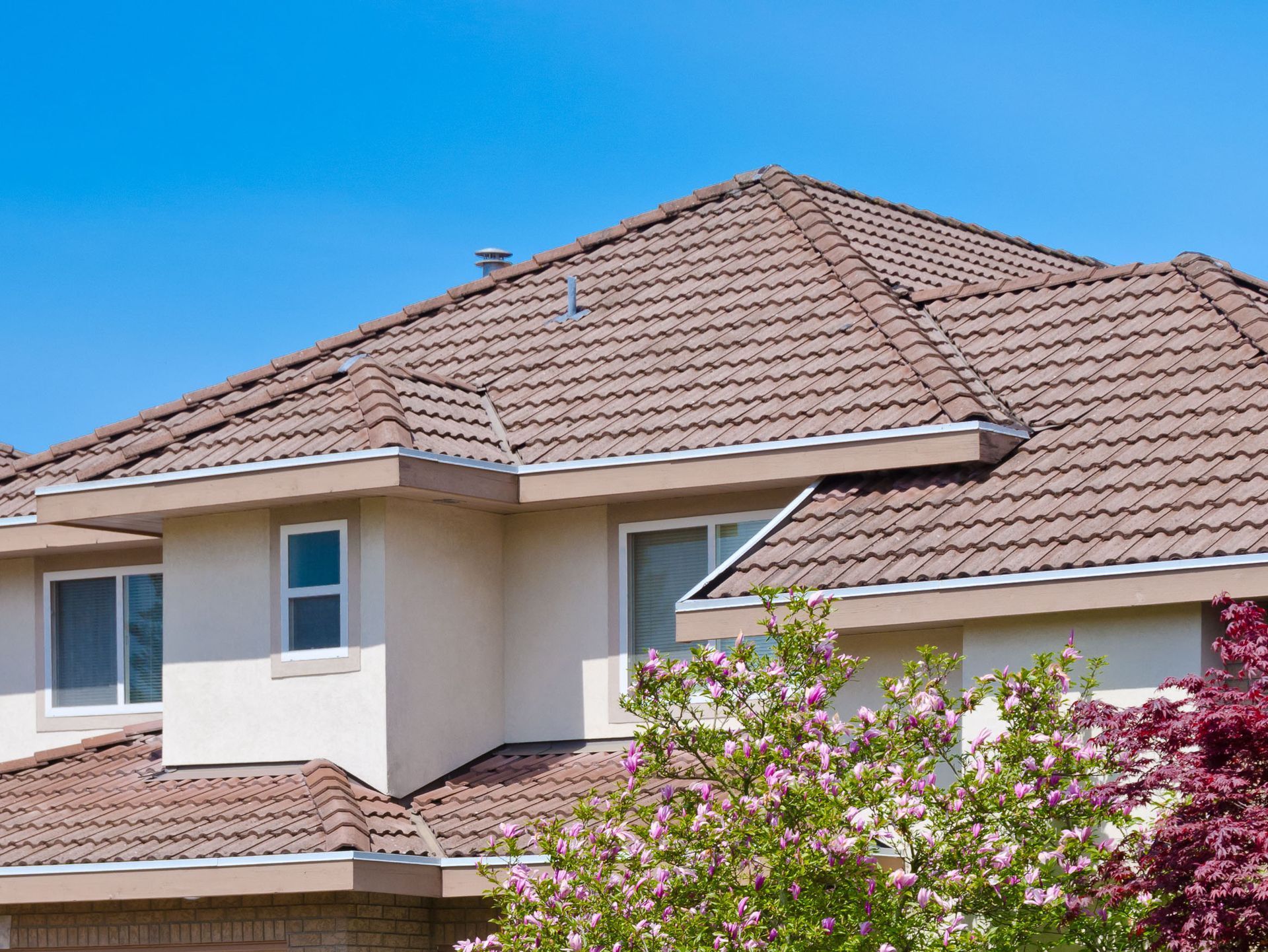 Two-story house with a brown tile roof and tan walls under a blue sky. Pink and red bushes in the foreground.