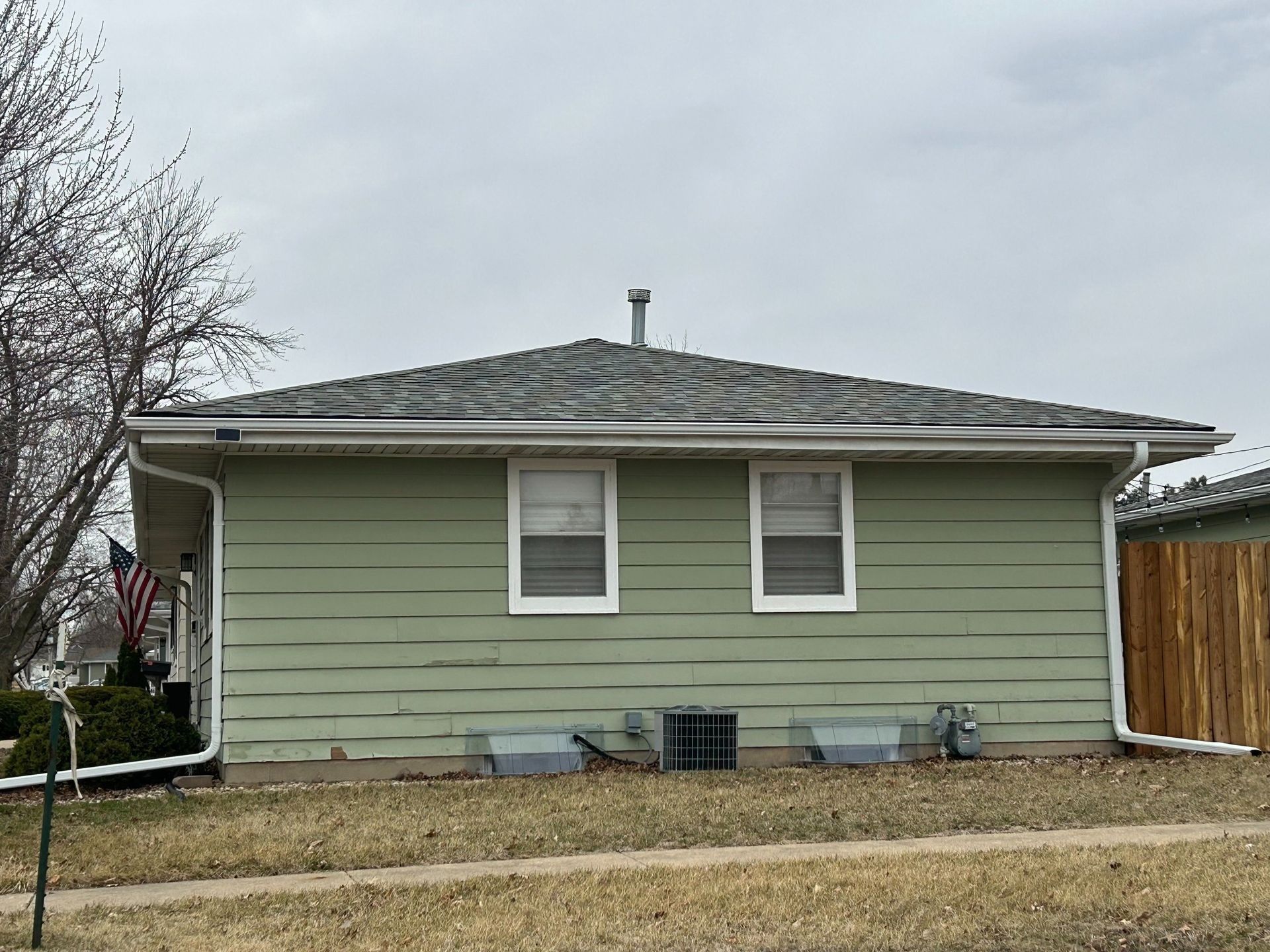 Green house with two windows, a chimney, and a gray roof under a cloudy sky.