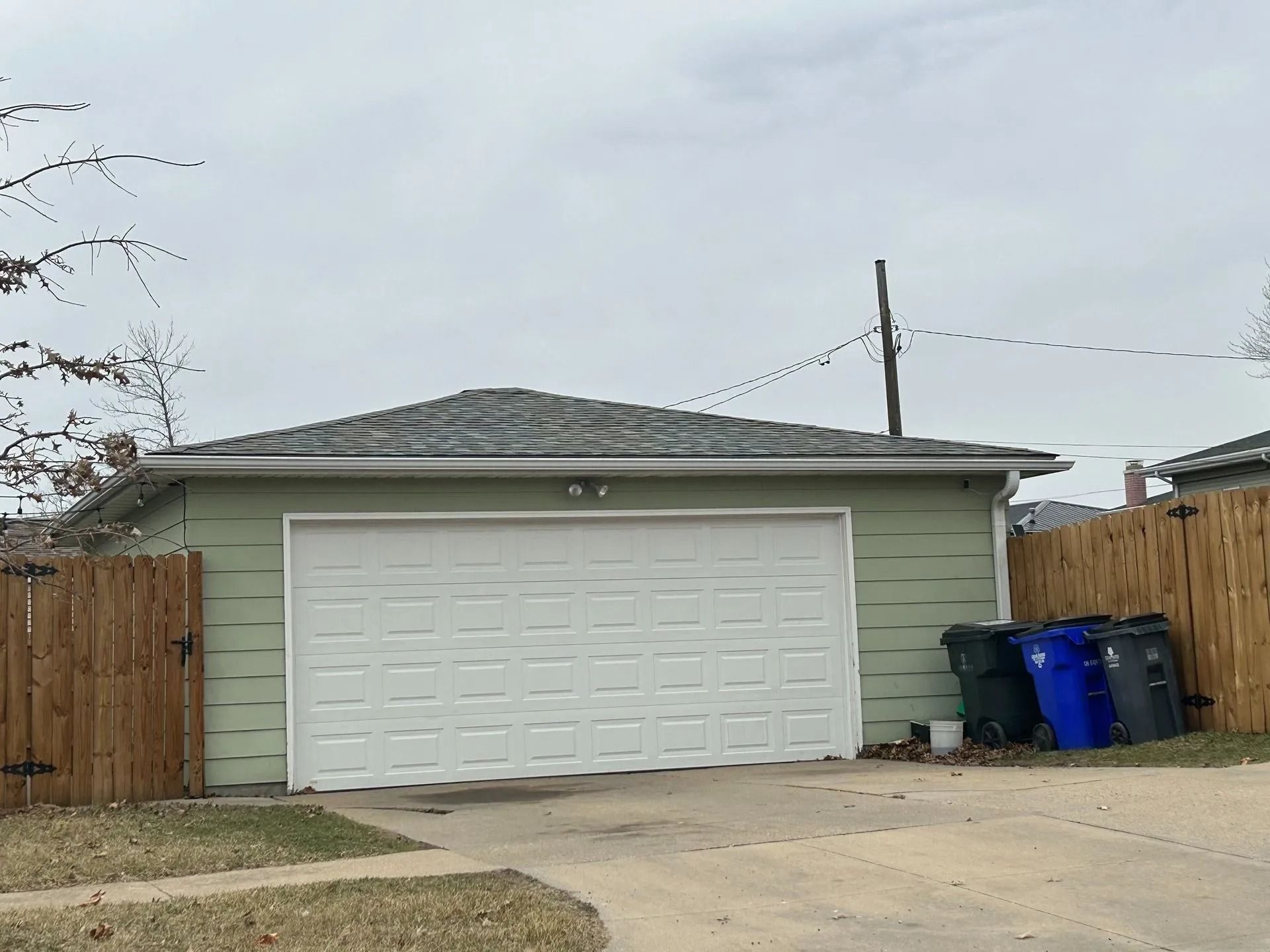 Green garage with a white door, brown fences, and trash bins. Overcast sky.