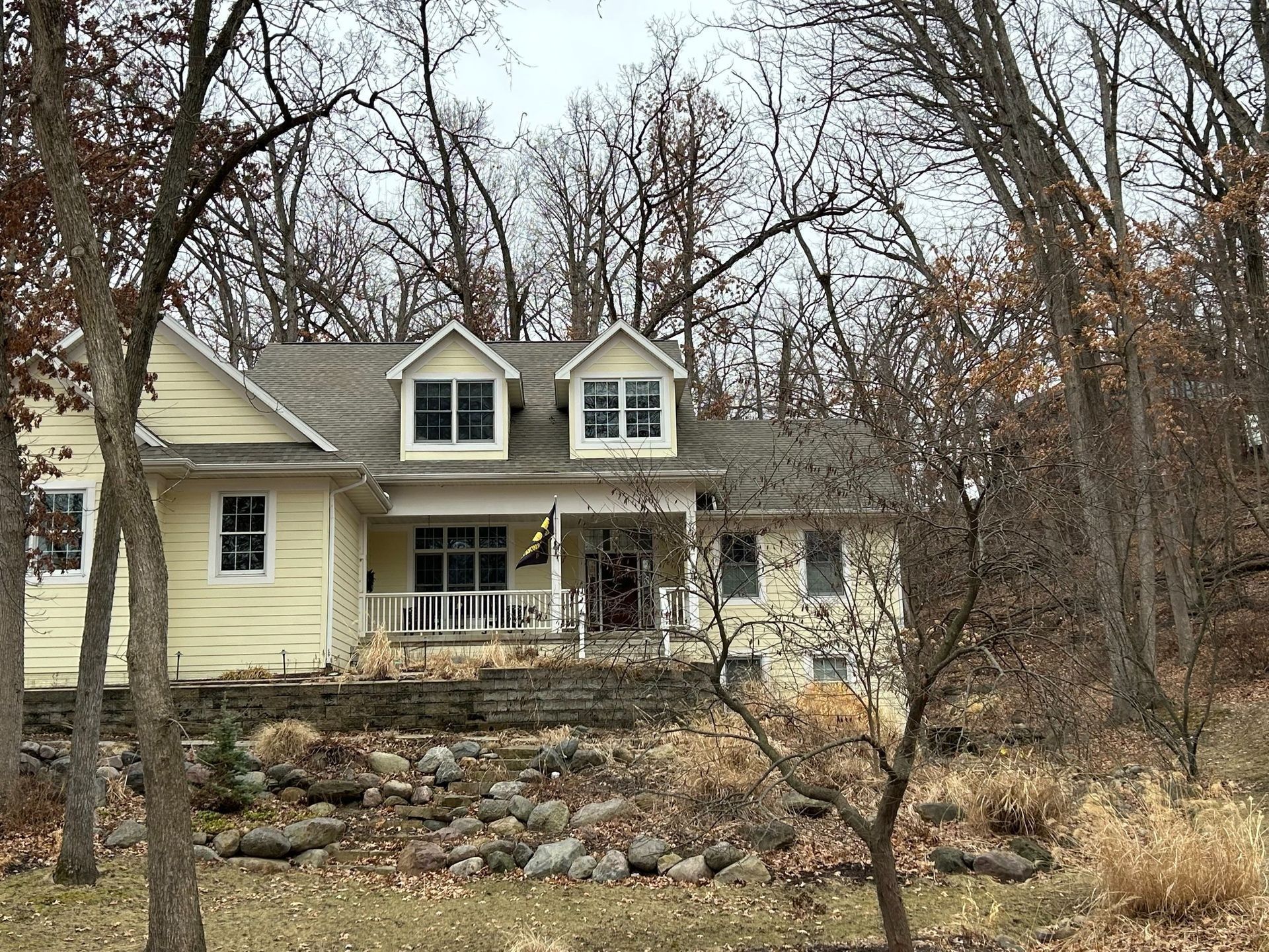 Yellow house with a porch and two dormers, set on a rocky hillside amongst bare trees.