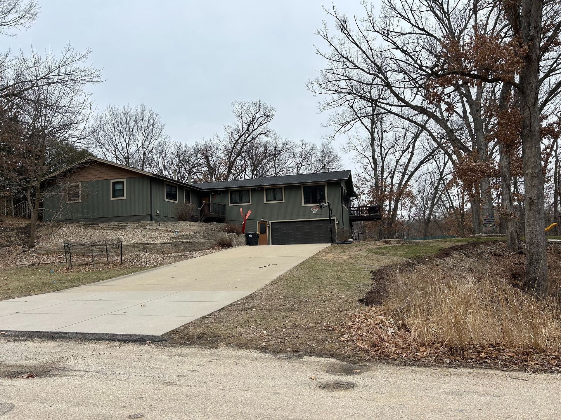 Green house with attached garage, long driveway, surrounded by bare trees, cloudy sky.