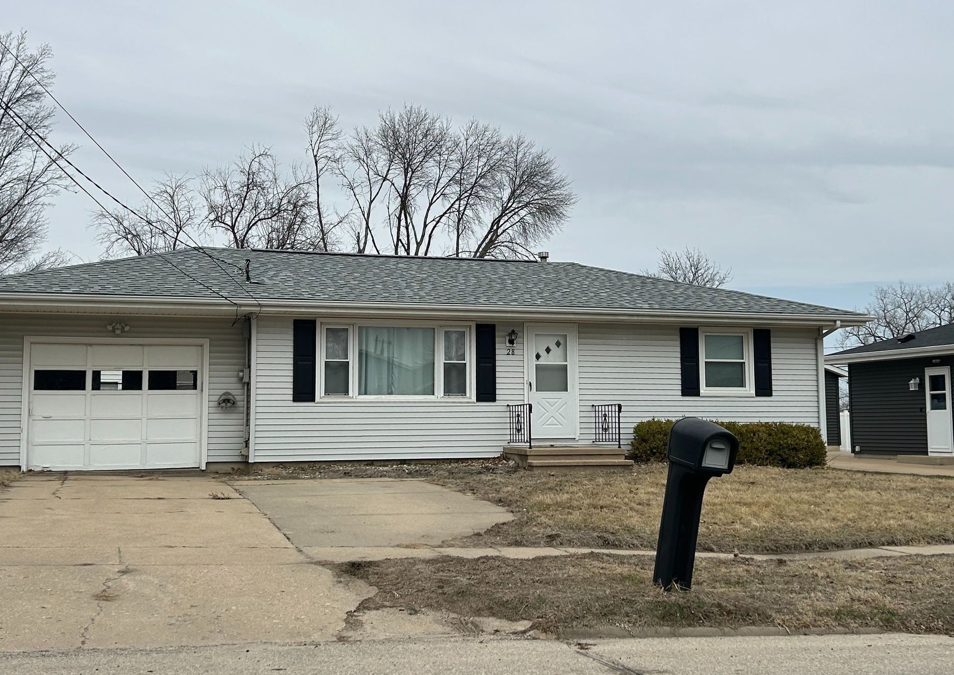 Ranch-style house with garage, faded white siding, black shutters, and mailbox on a cloudy day.