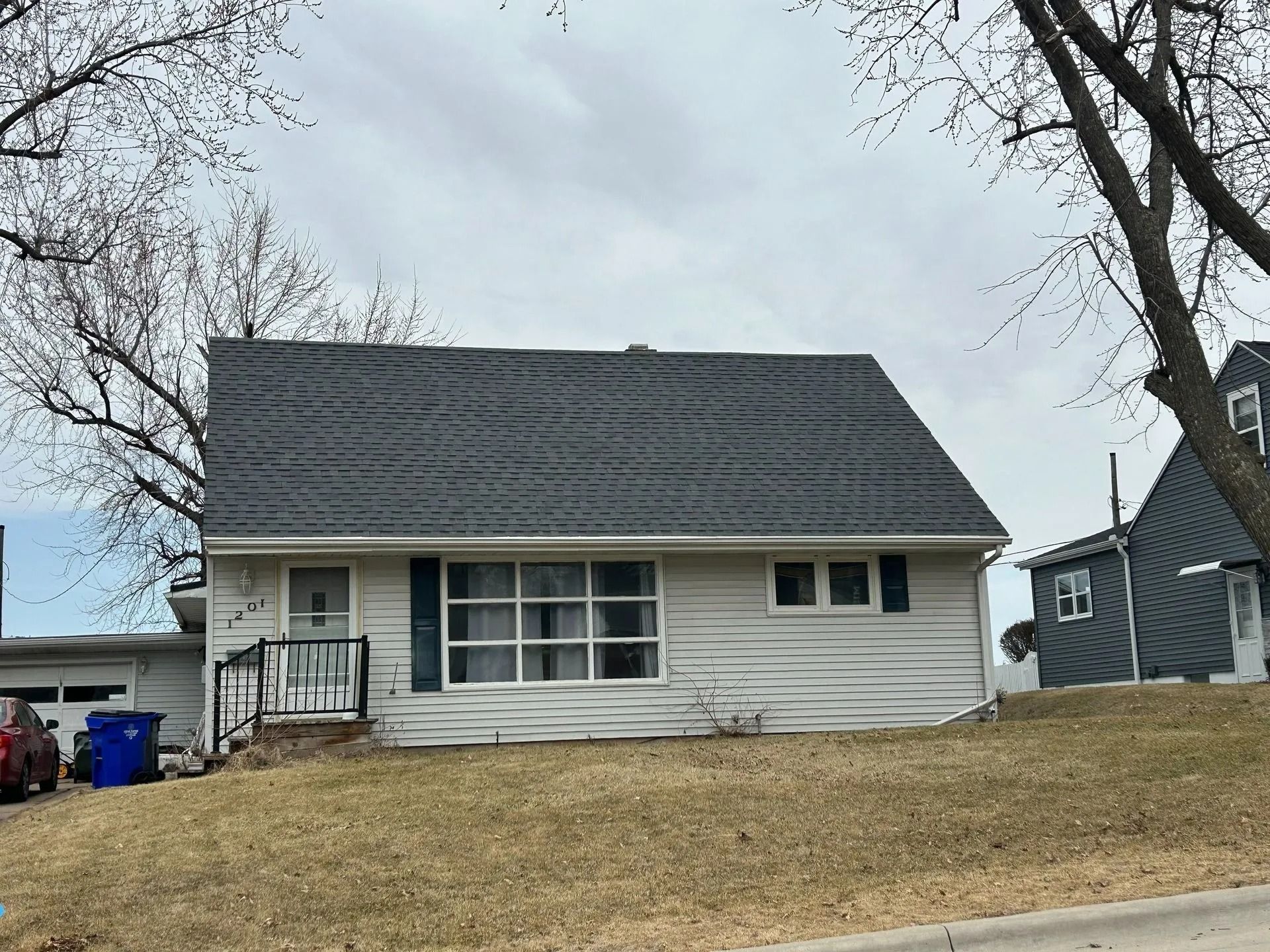 White house with dark gray roof, green shutters, and brown lawn under a cloudy sky.