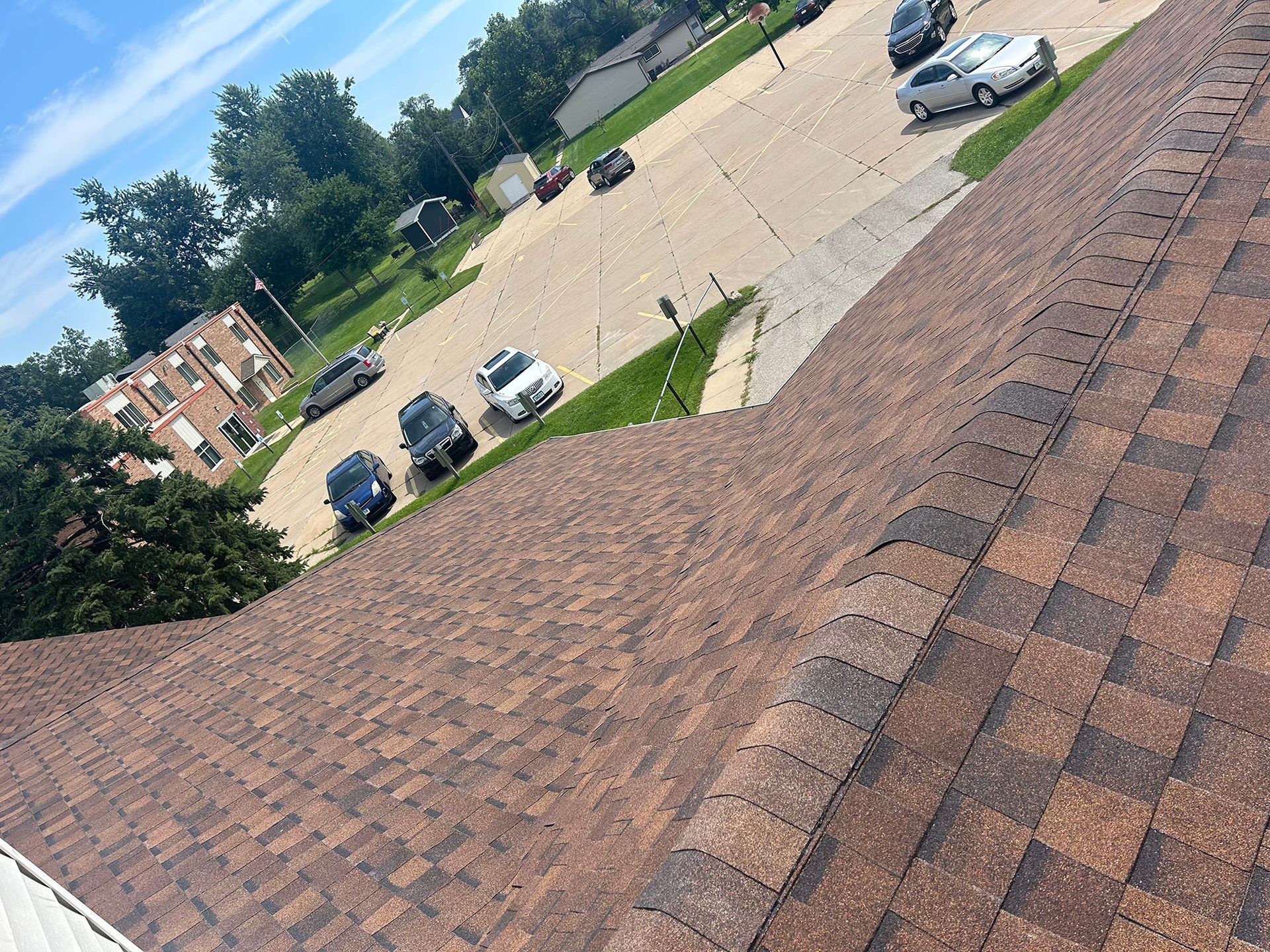 Brown asphalt shingle roof, angled view overlooking a street with parked cars and trees under a blue sky.