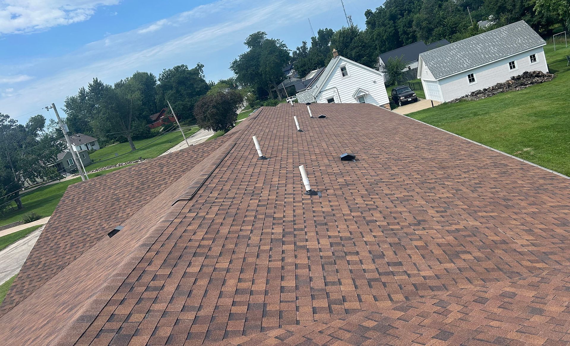Brown shingle roof of a house with white vent pipes. Green grass and trees in the background.