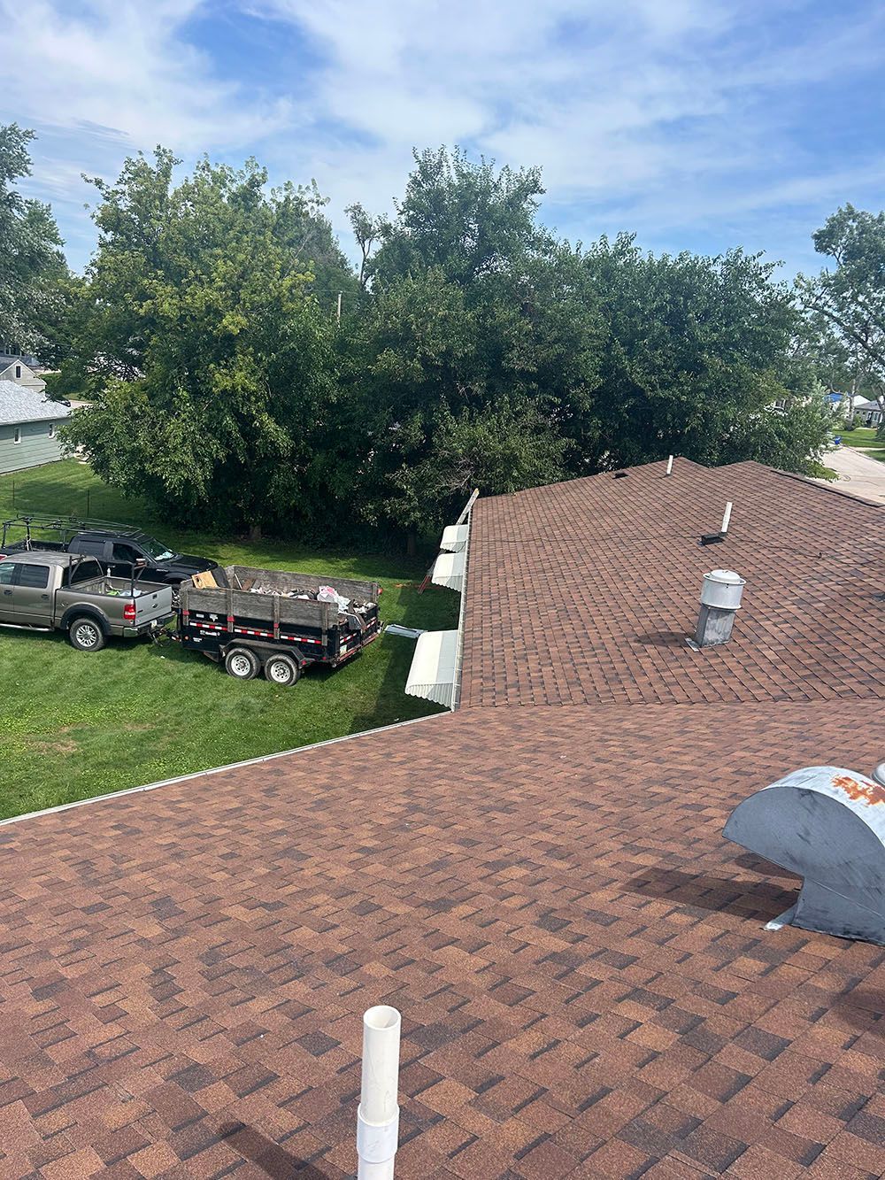 Brown shingled roof with a truck and trailer on the lawn; trees in the background under a blue sky.