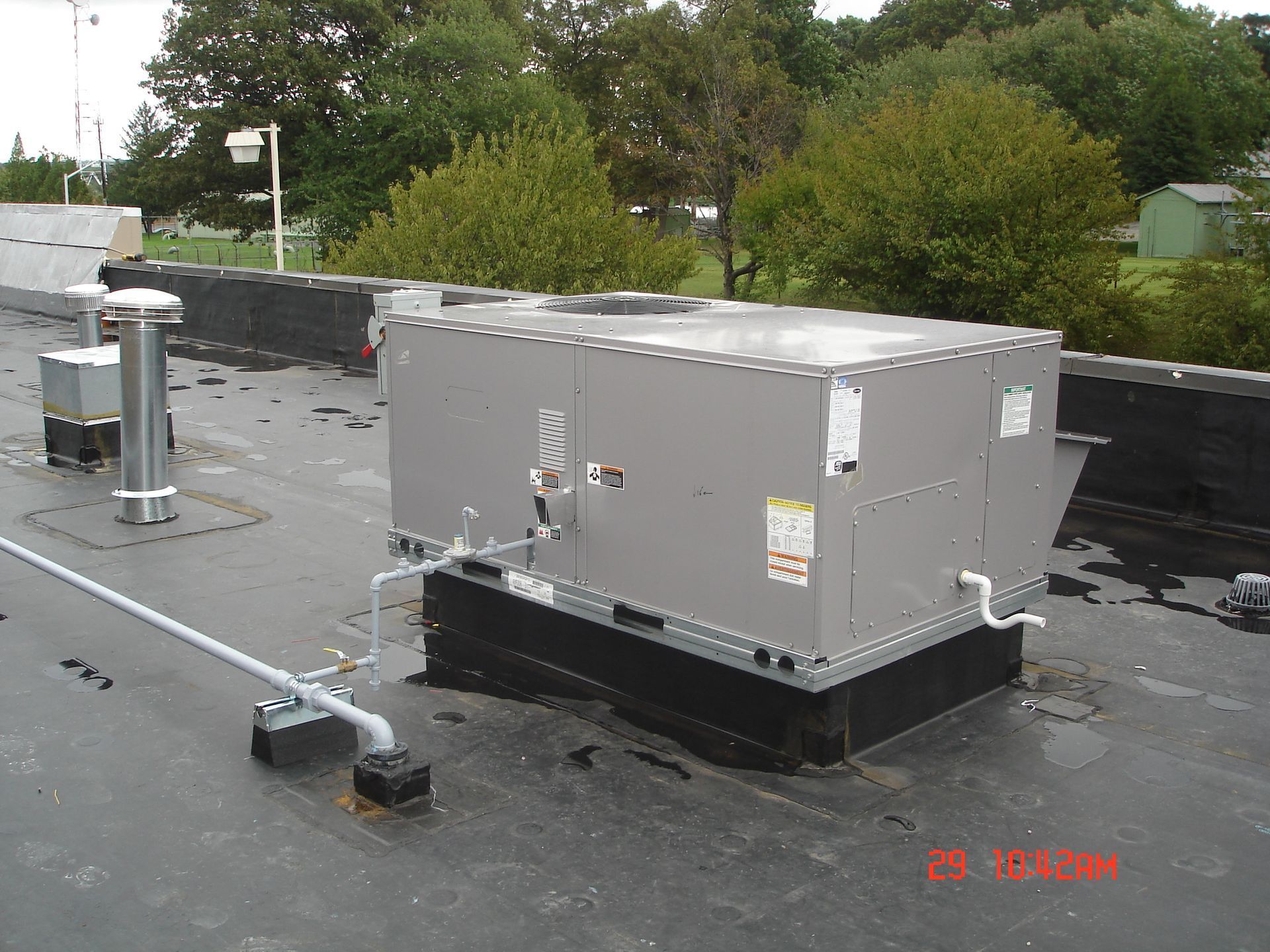Rooftop HVAC units and ductwork on a flat roof with trees in the background.