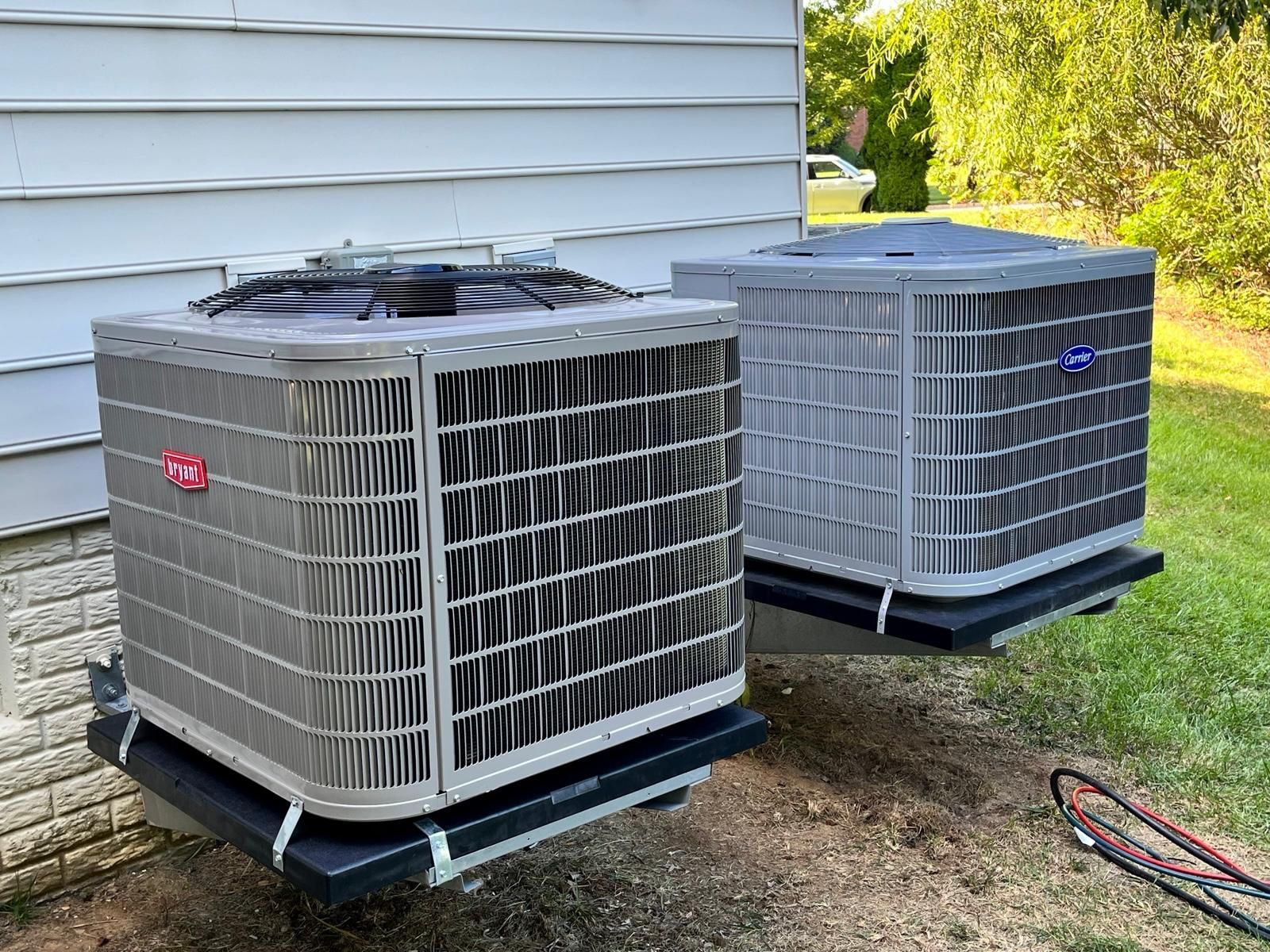 Two outdoor air conditioning units beside a house, one gray and one blue-gray, on a gravel pad.