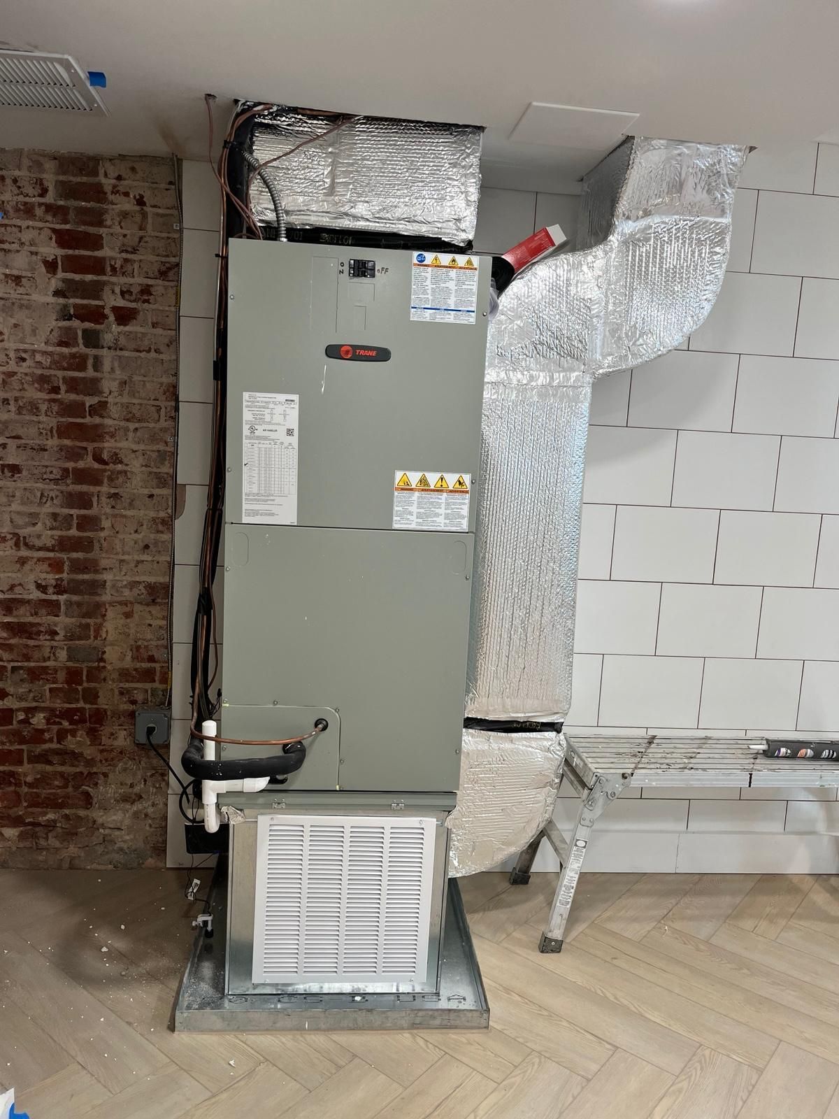 HVAC furnace unit with insulated ductwork in a partially finished basement, beside a brick wall and cinder blocks