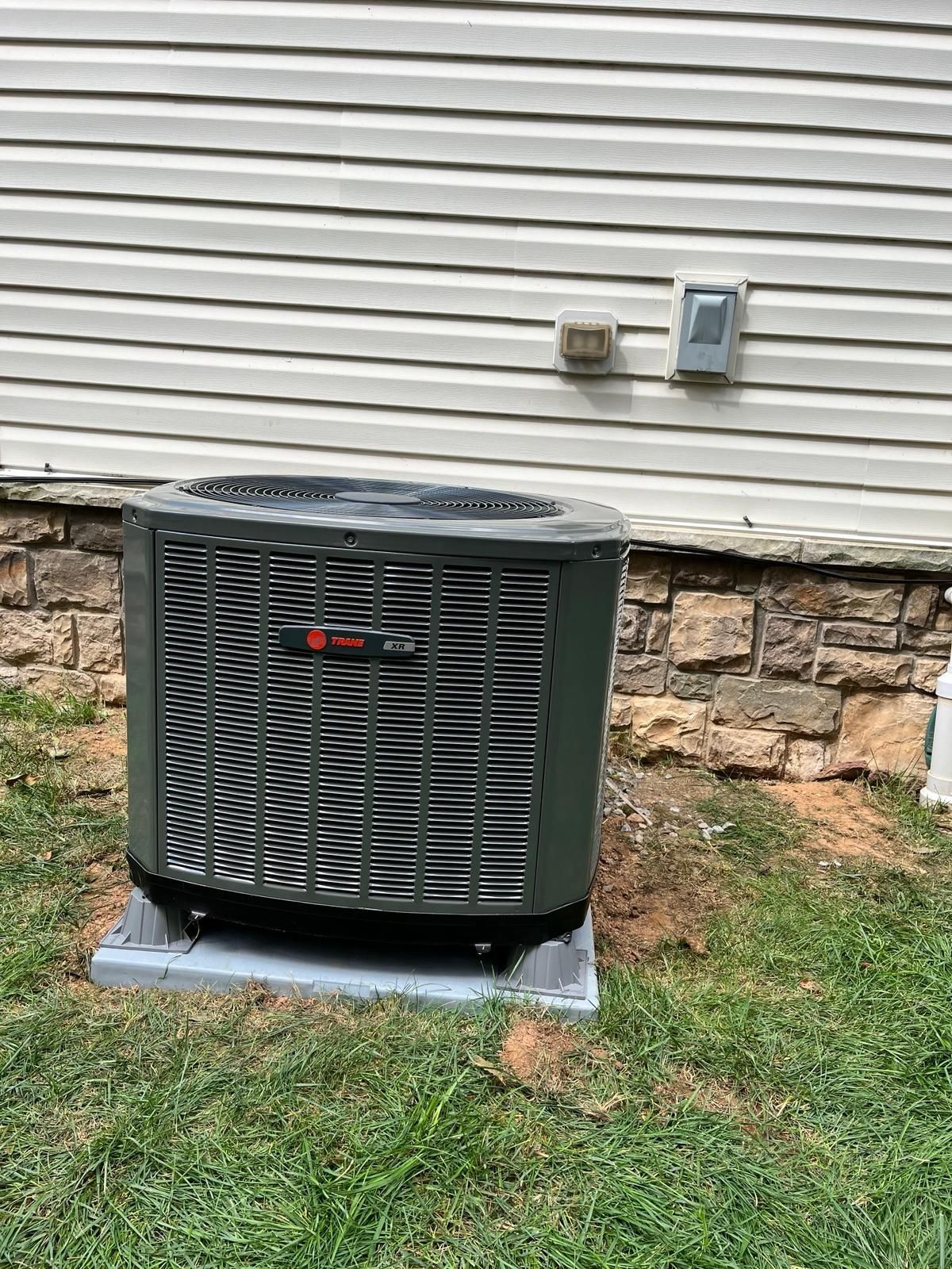 Outdoor air conditioning unit beside a house wall, on a concrete pad with grass around it.