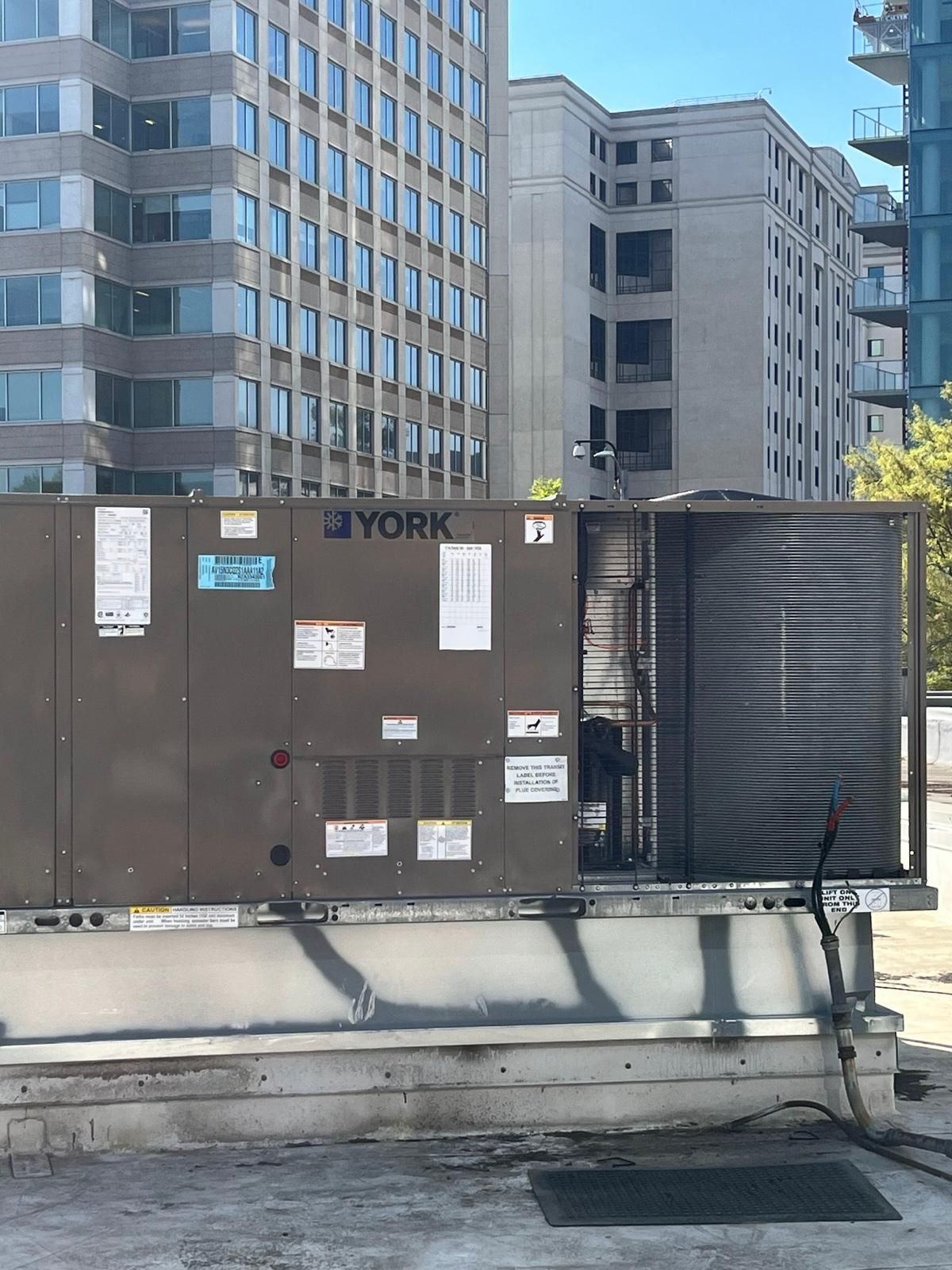 Rooftop HVAC unit with vents and labels, set against tall apartment buildings and blue sky.