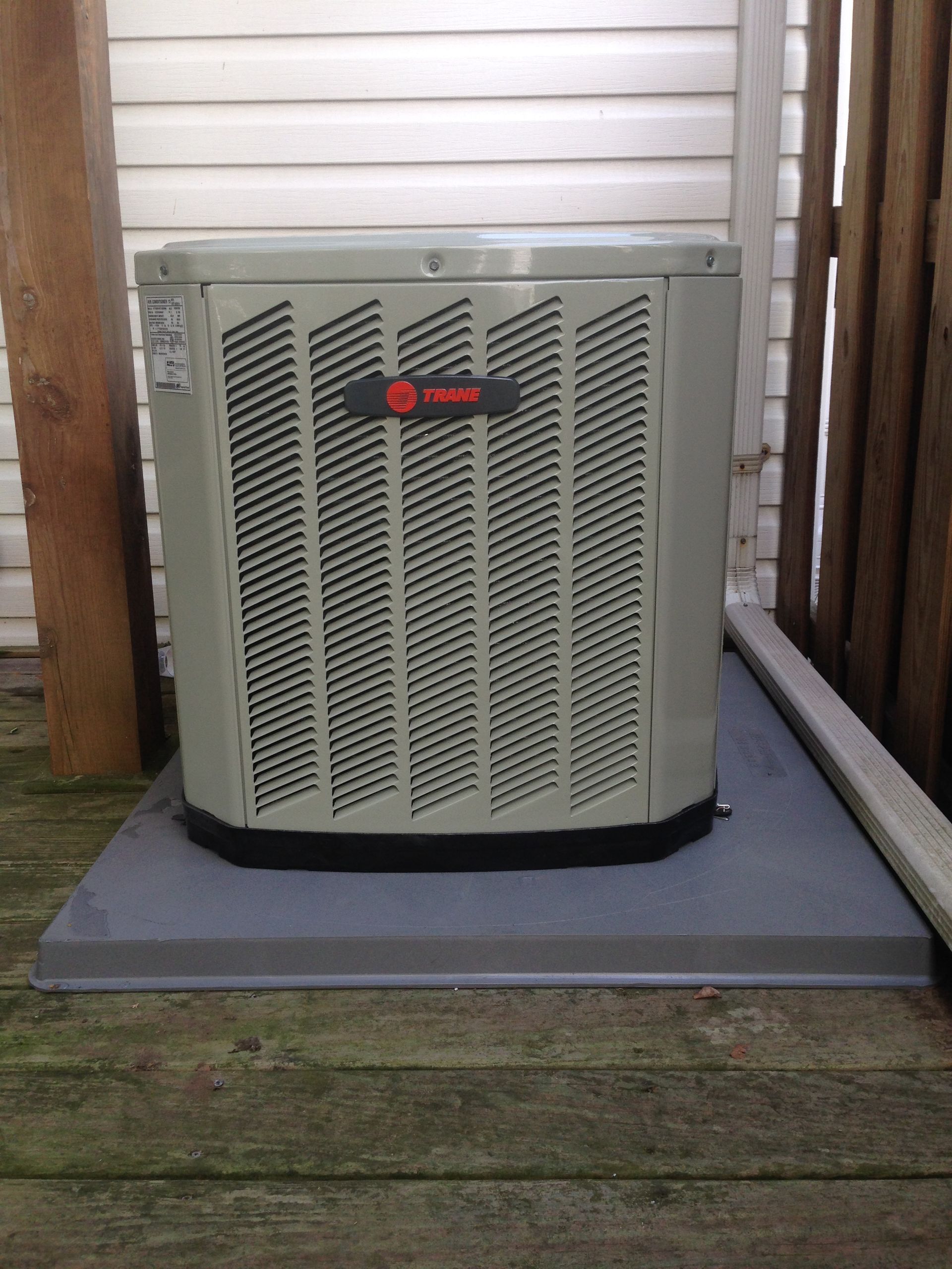 Outdoor HVAC unit on a gray platform beside a house wall and wooden fence