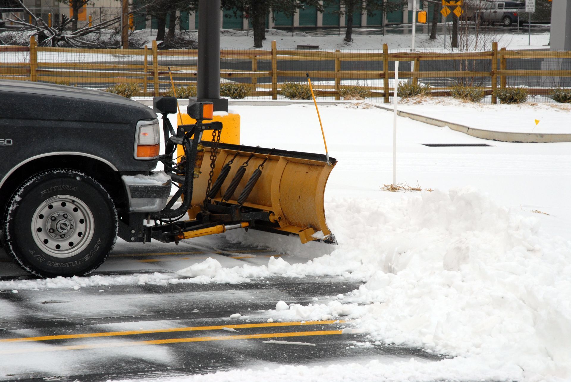 A black truck with a yellow snowplow clearing snow from a parking lot.