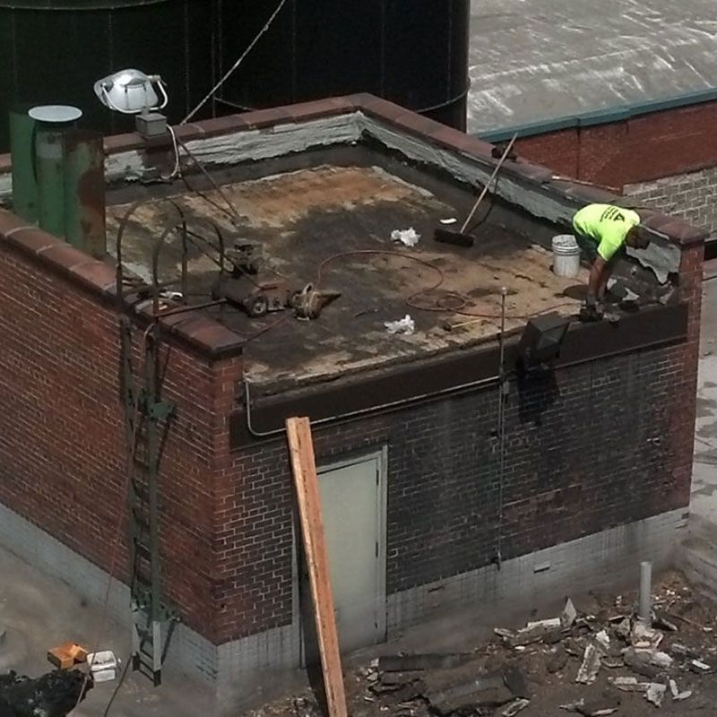 Roofer in neon vest on a brick building's flat roof, removing old materials.