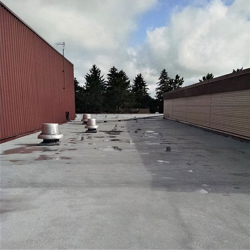 Gray flat roof with vents, red building on the left, brown building on the right, trees and cloudy sky in background.