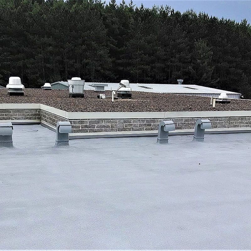 Gray flat roof with various vents, topped with dark pebbles. Building in background, trees in the distance.