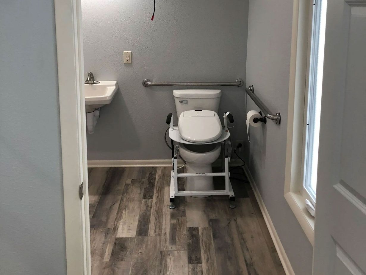 Accessible bathroom with raised toilet, grab bars, and sink. Gray walls, wood-look floor.