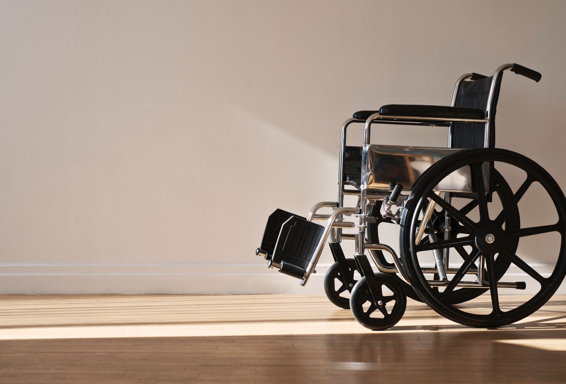 Wheelchair on wood floor against white wall, illuminated by sunlight.