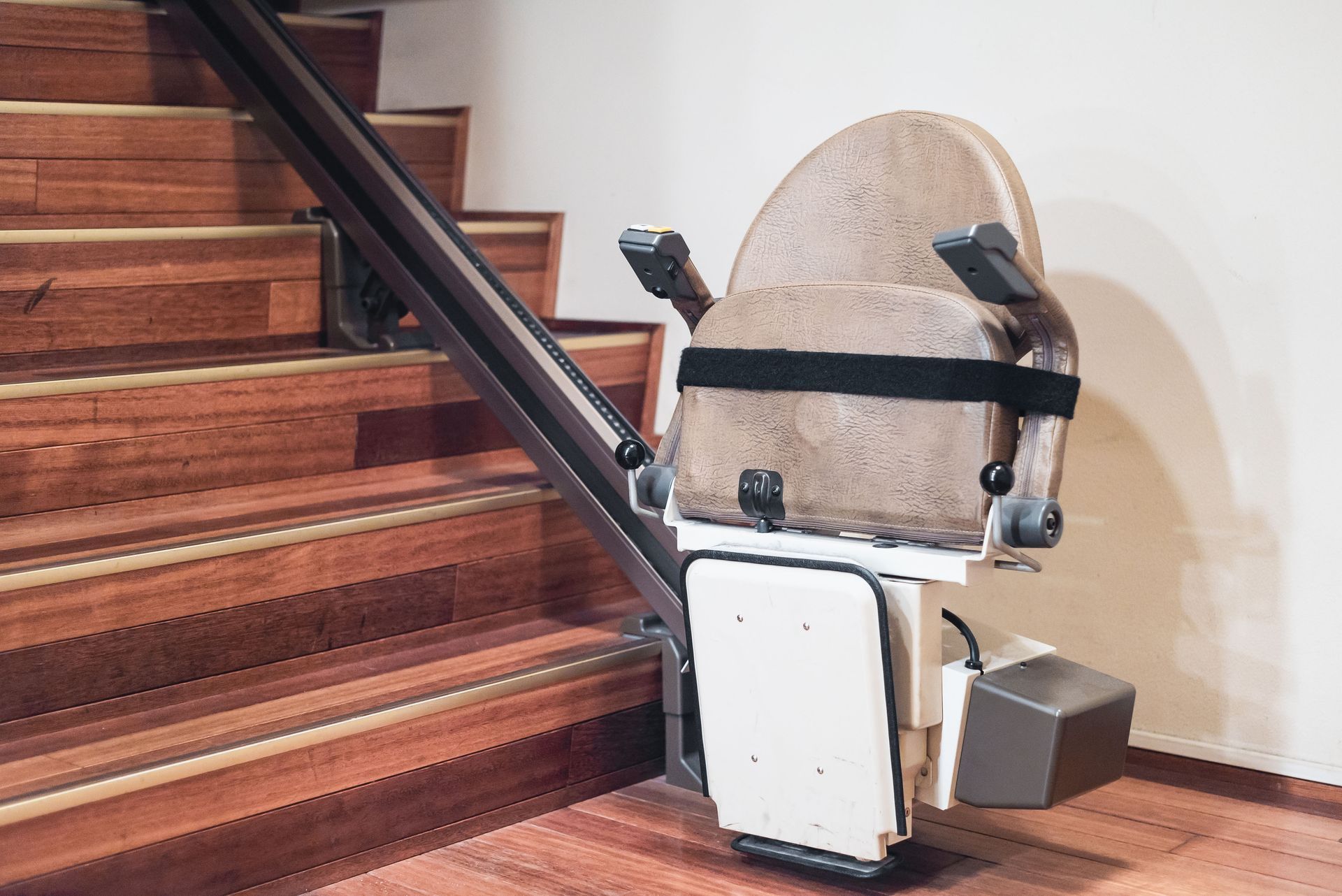 Stair lift installed on wooden staircase. Tan seat with armrests, black safety belt, and metal rail.