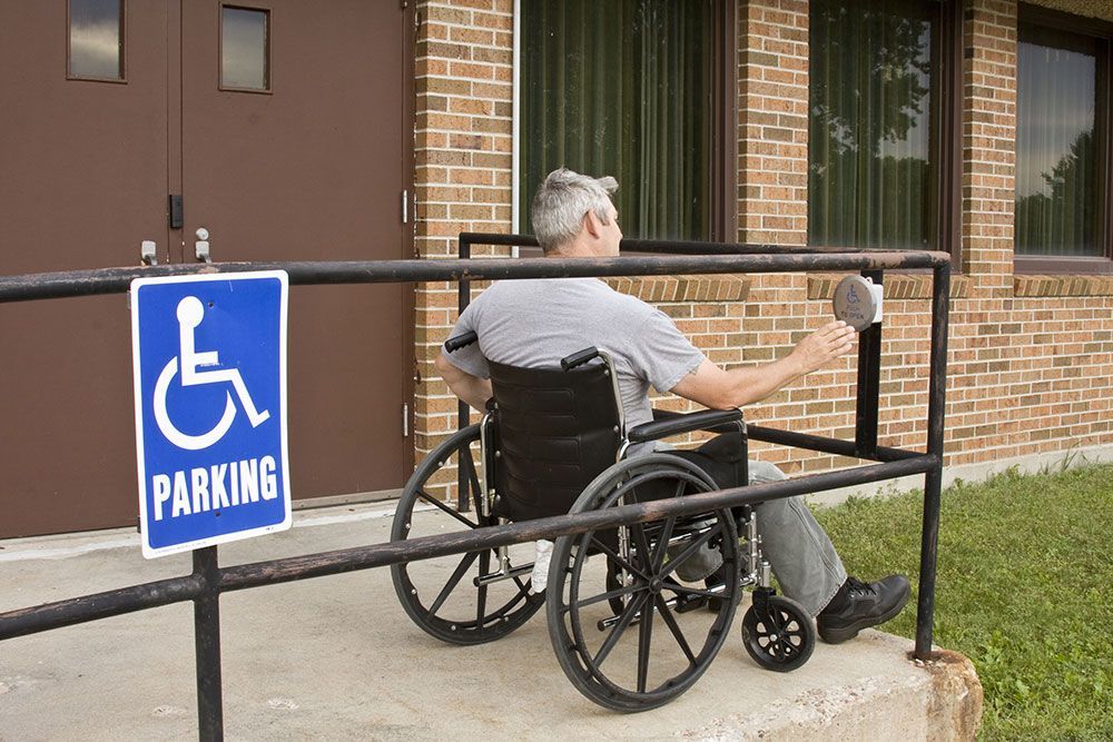 Man in wheelchair uses a ramp to enter a building with a handicap parking sign.