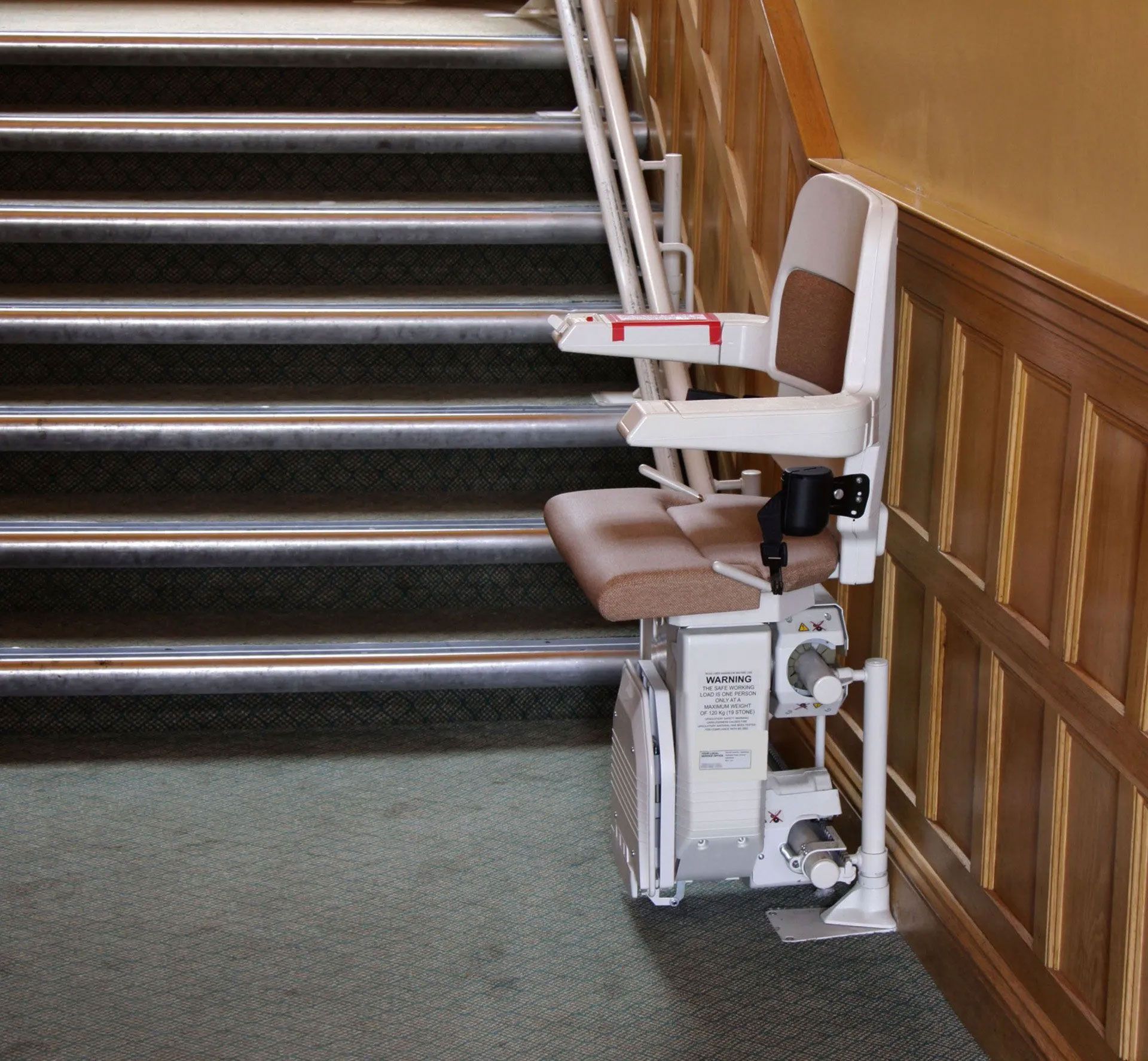 Stair lift installed on stairs. White chair, brown cushion, next to wood paneling.