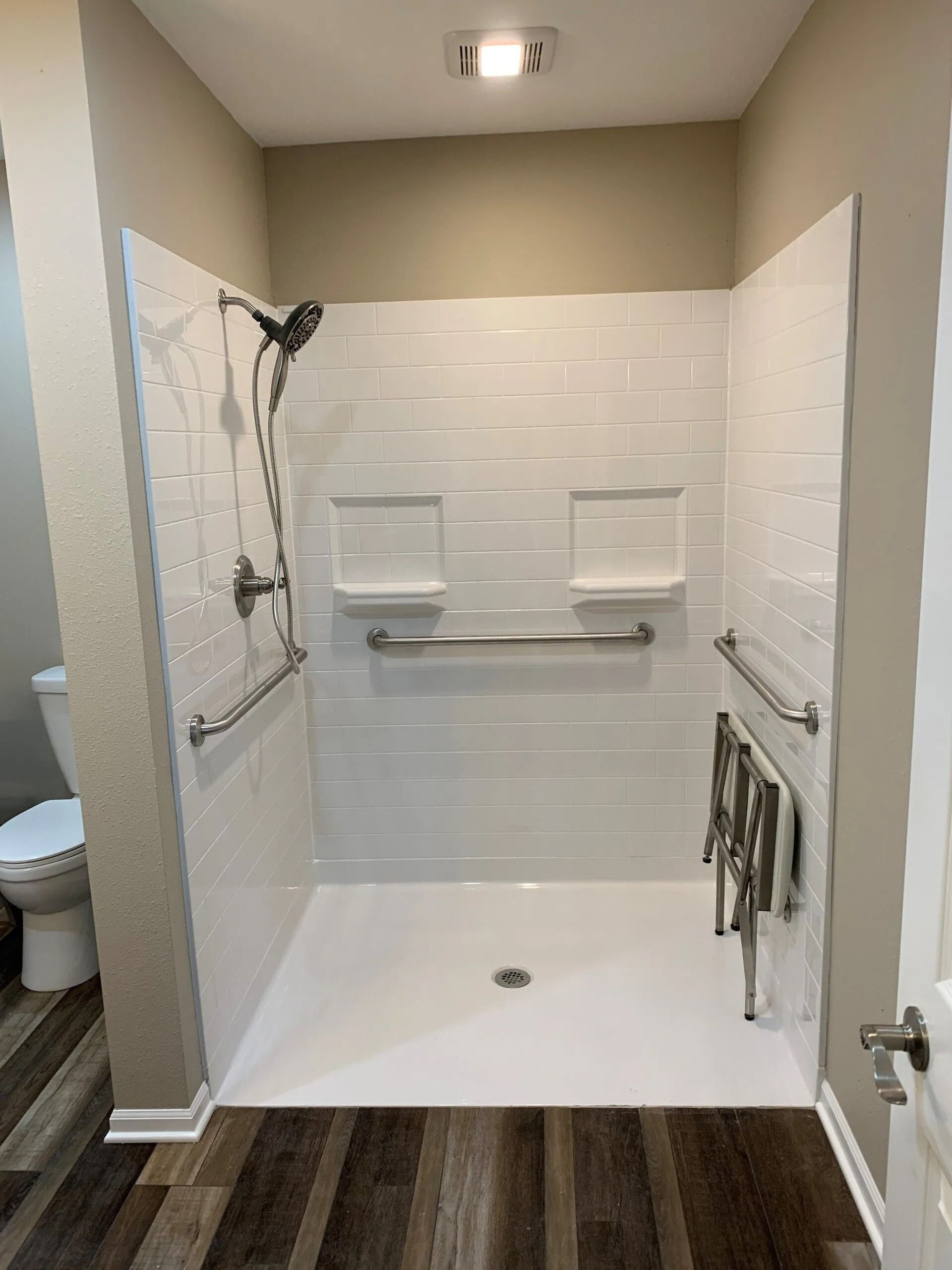Accessible shower with grab bars and built-in shelves; white tiled walls, brown floor, and a toilet to the left.