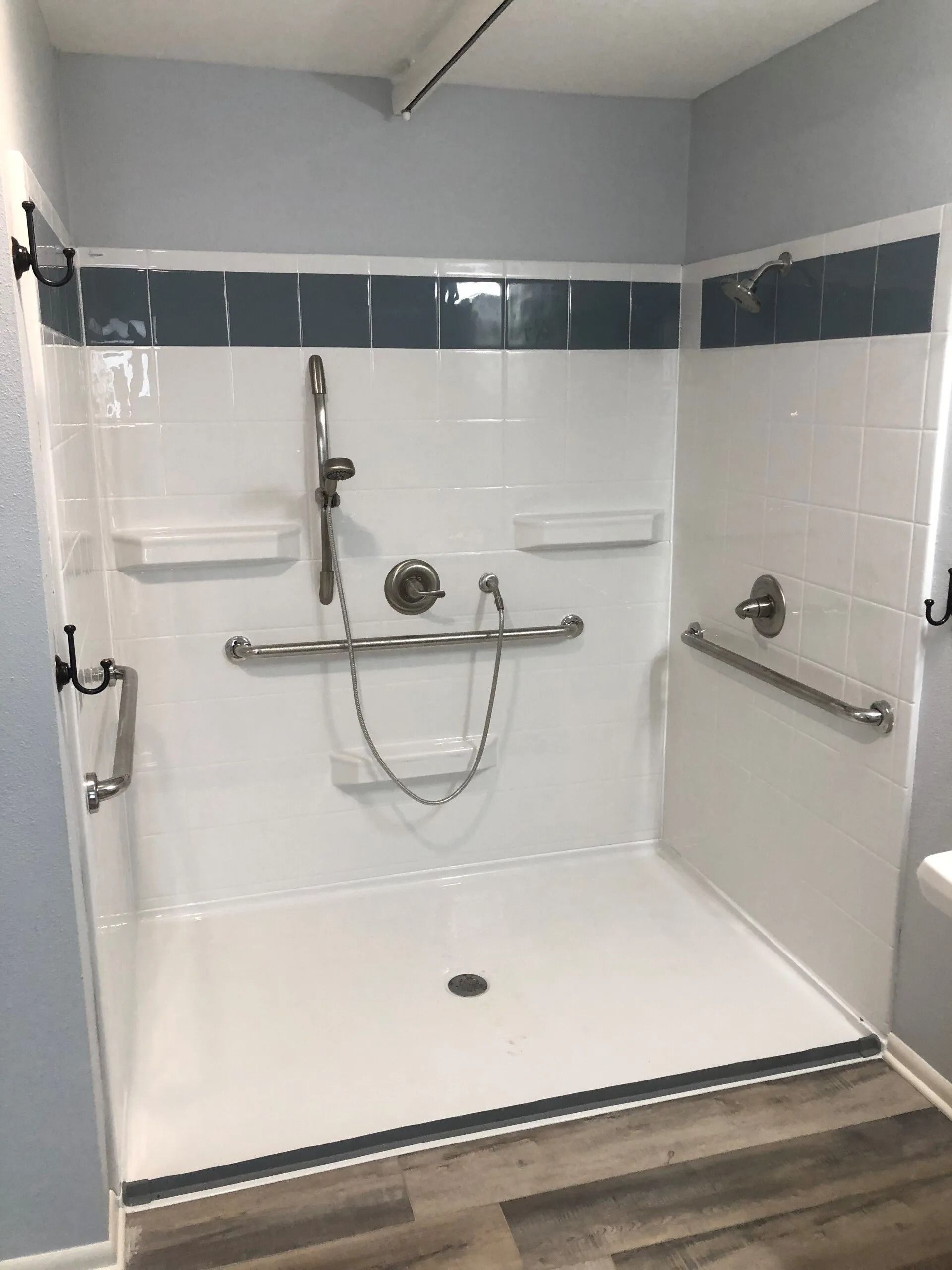 Accessible shower with grab bars, shower head, white walls, gray and dark blue trim, and wooden floor.
