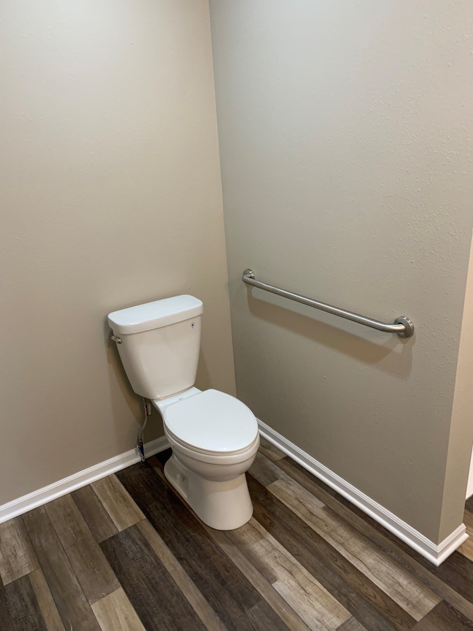 Toilet in a corner with a grab bar on the wall. Brown and white wood-look flooring. Light beige walls.