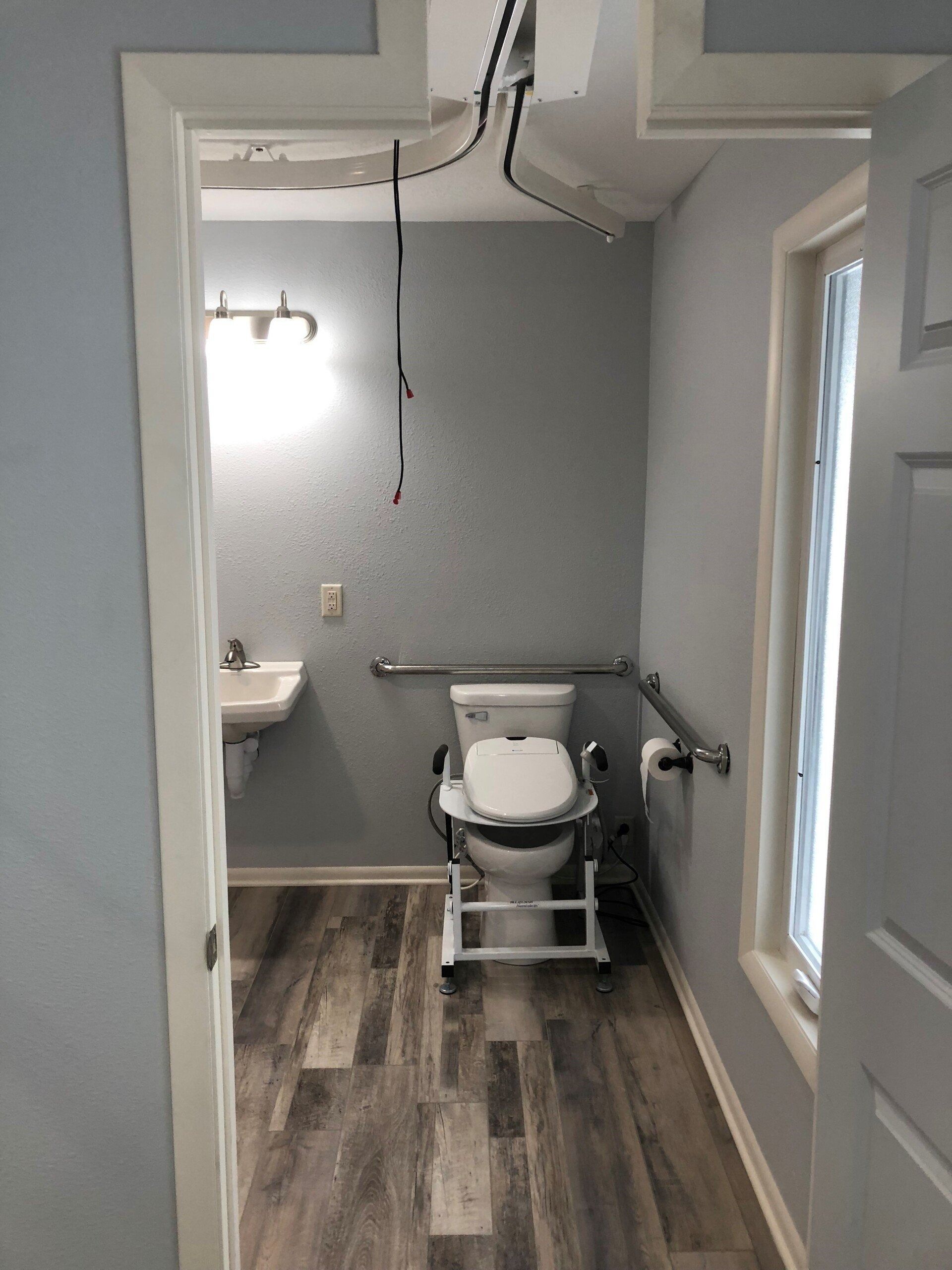 ADA-accessible bathroom with a raised toilet, grab bars, and a small window. Gray walls, wood-look floor.