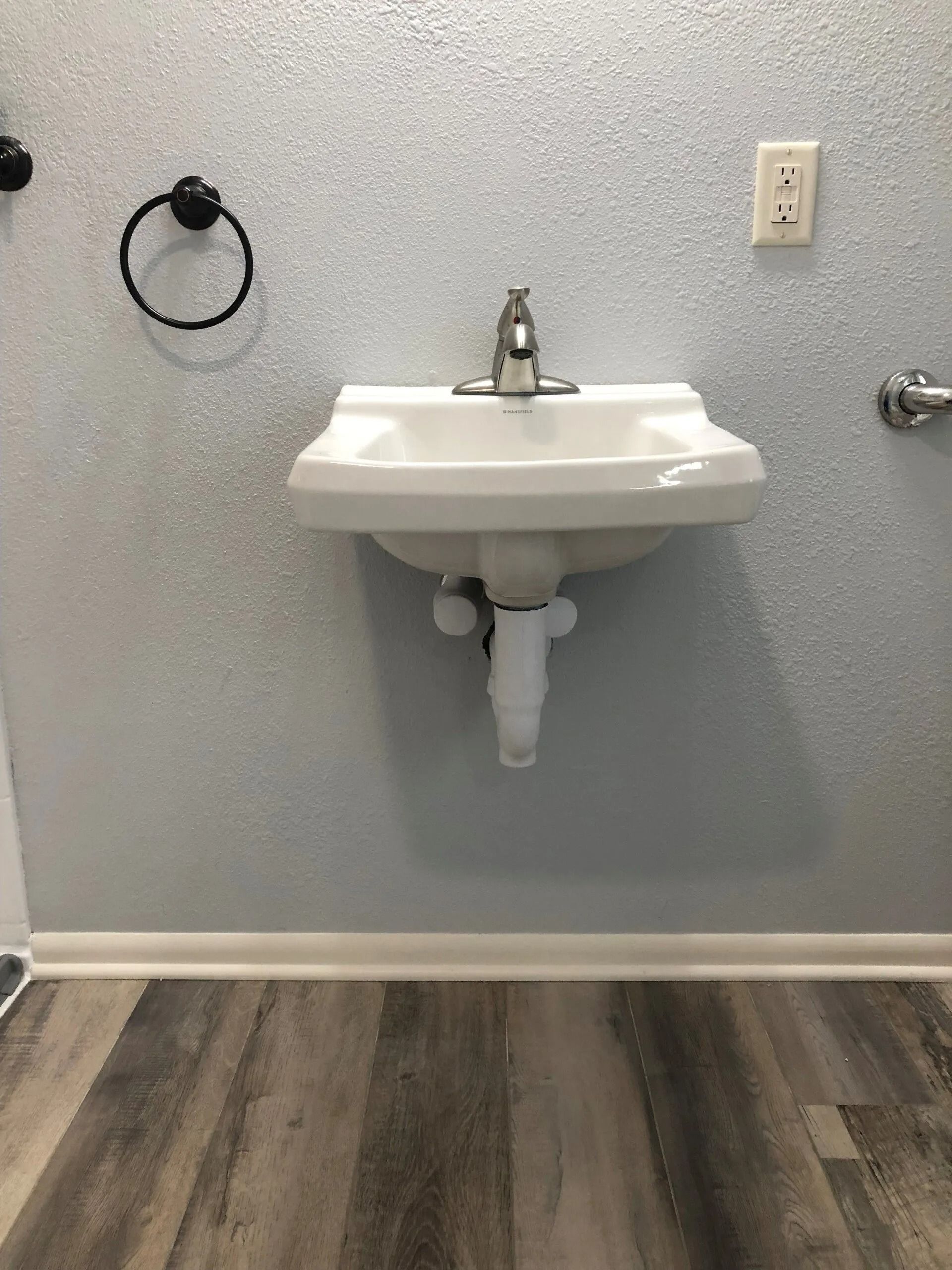 White wall-mounted sink with chrome faucet, black towel ring, and white wall outlet in bathroom.