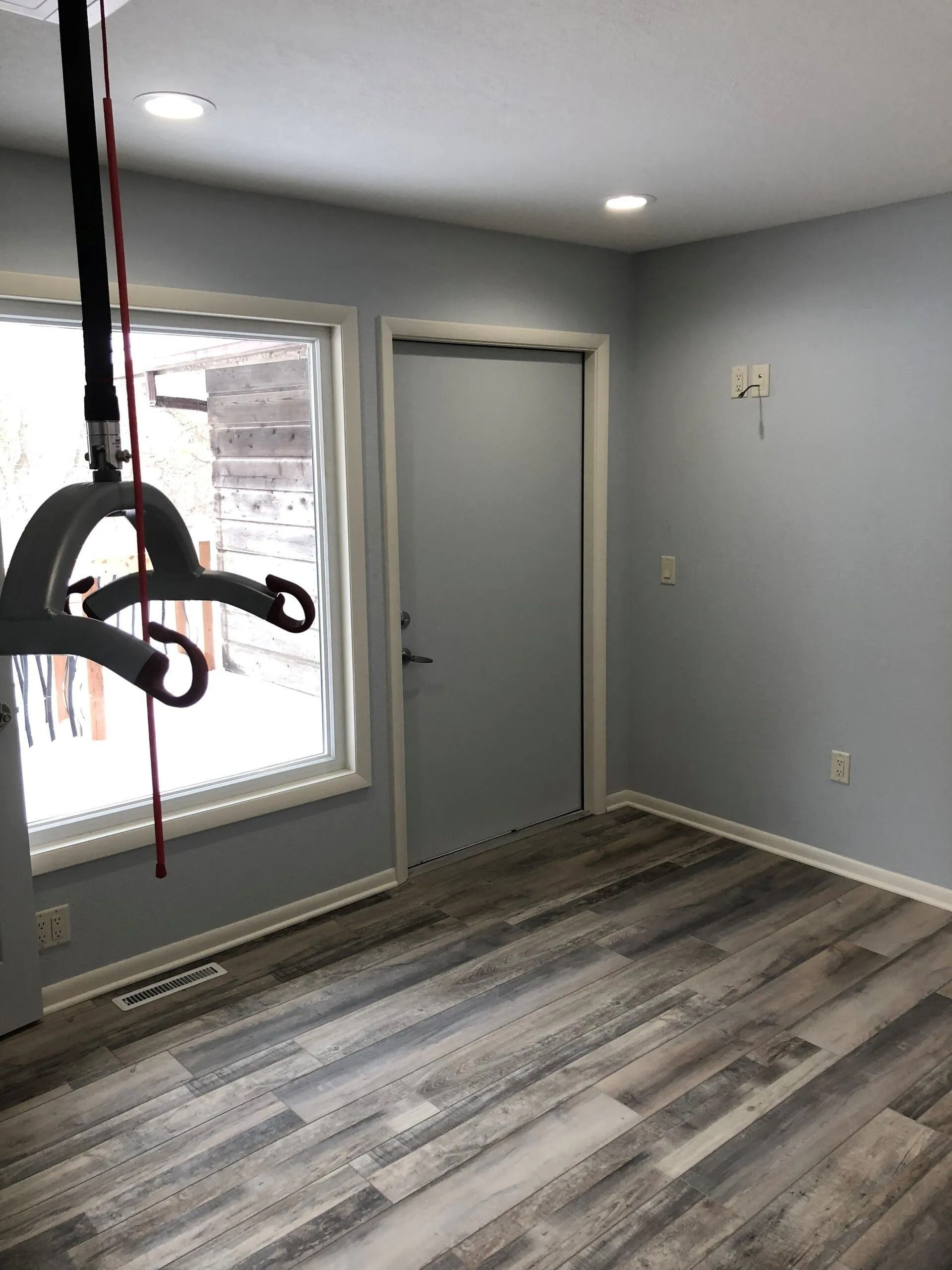 Empty room with light gray walls, wood-look floor, window, and gray door. Ceiling lights and lift apparatus.